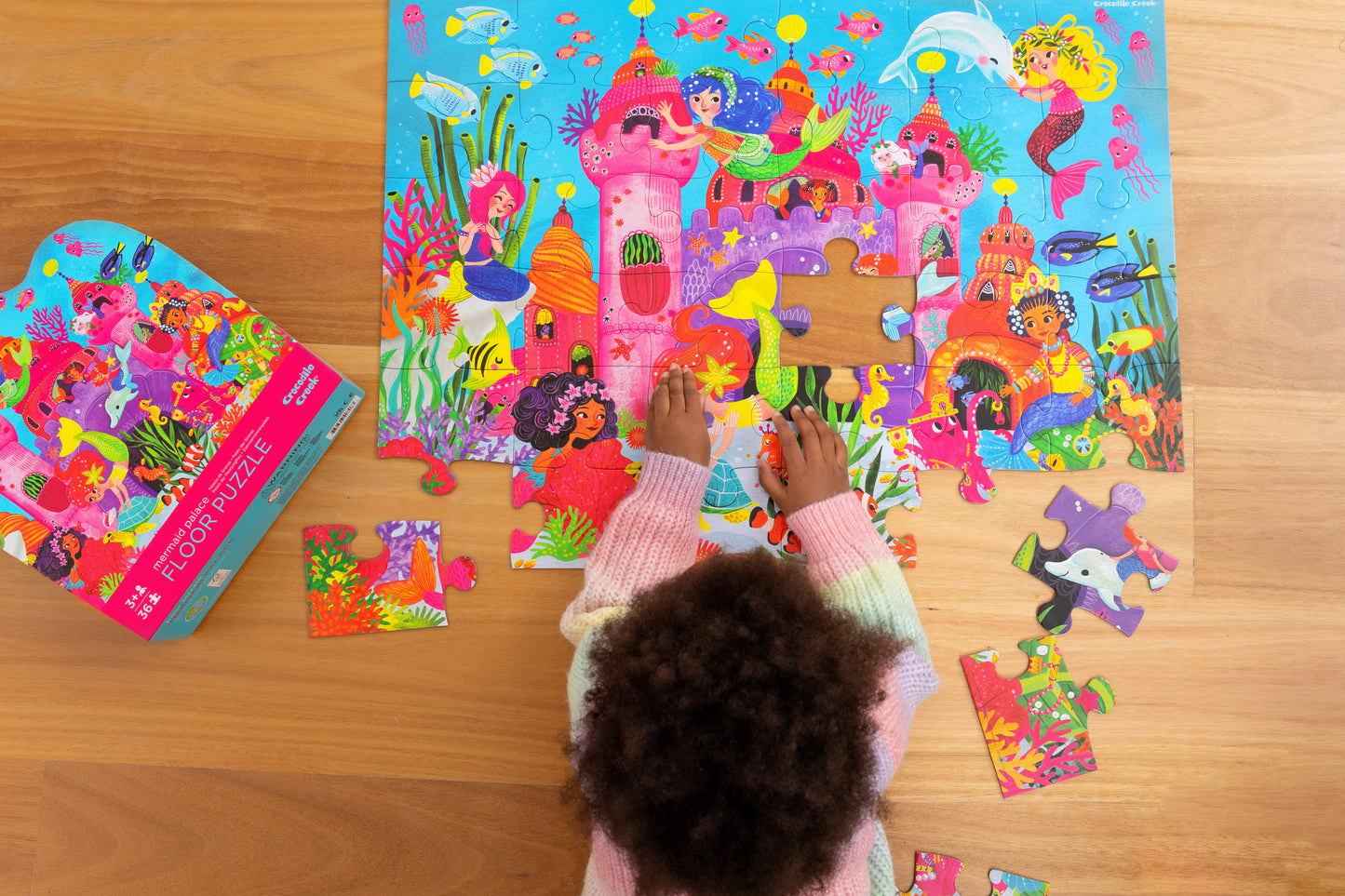 A child is assembling a colourful floor puzzle featuring mermaids, dolphins, and treasure chests in a room.