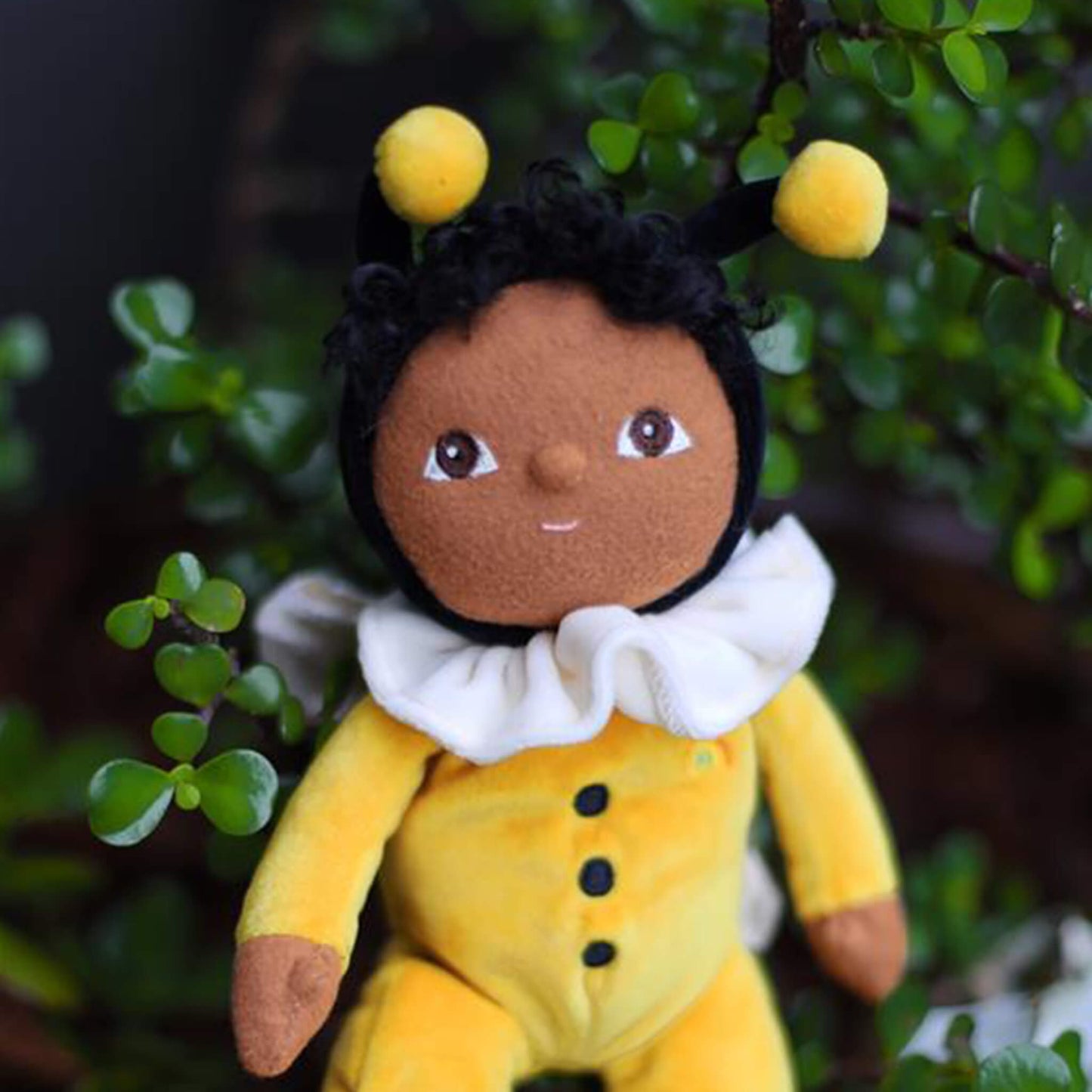 Toy bee character with a yellow body and black hair, standing among green leaves.