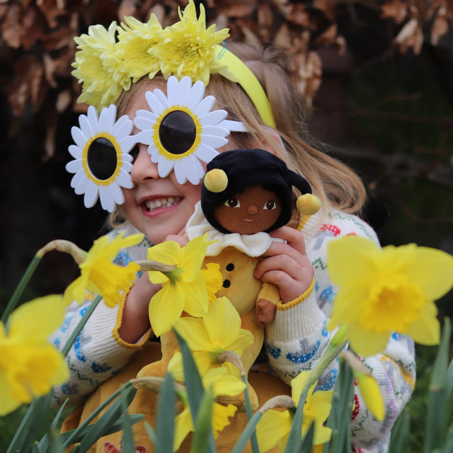 Child with floral headband and daffodils, holding a doll, surrounded by flowers.