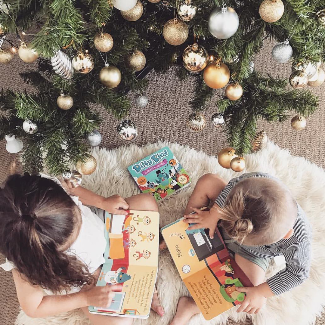 Two children reading books under a decorated Christmas tree.