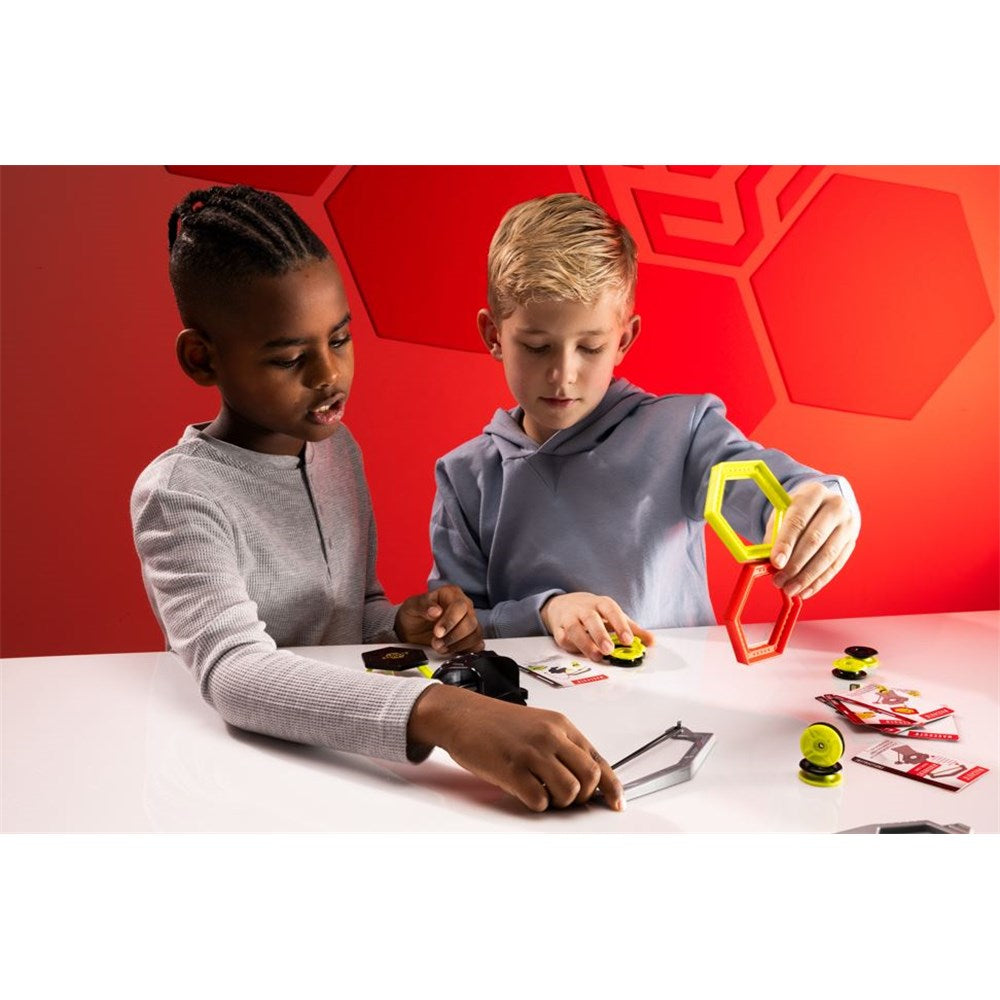 Two children playing with geometric shapes at a table against a red geometric-patterned wall.