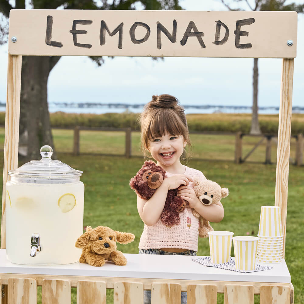 Child with stuffed animals behind a lemonade stand outdoors