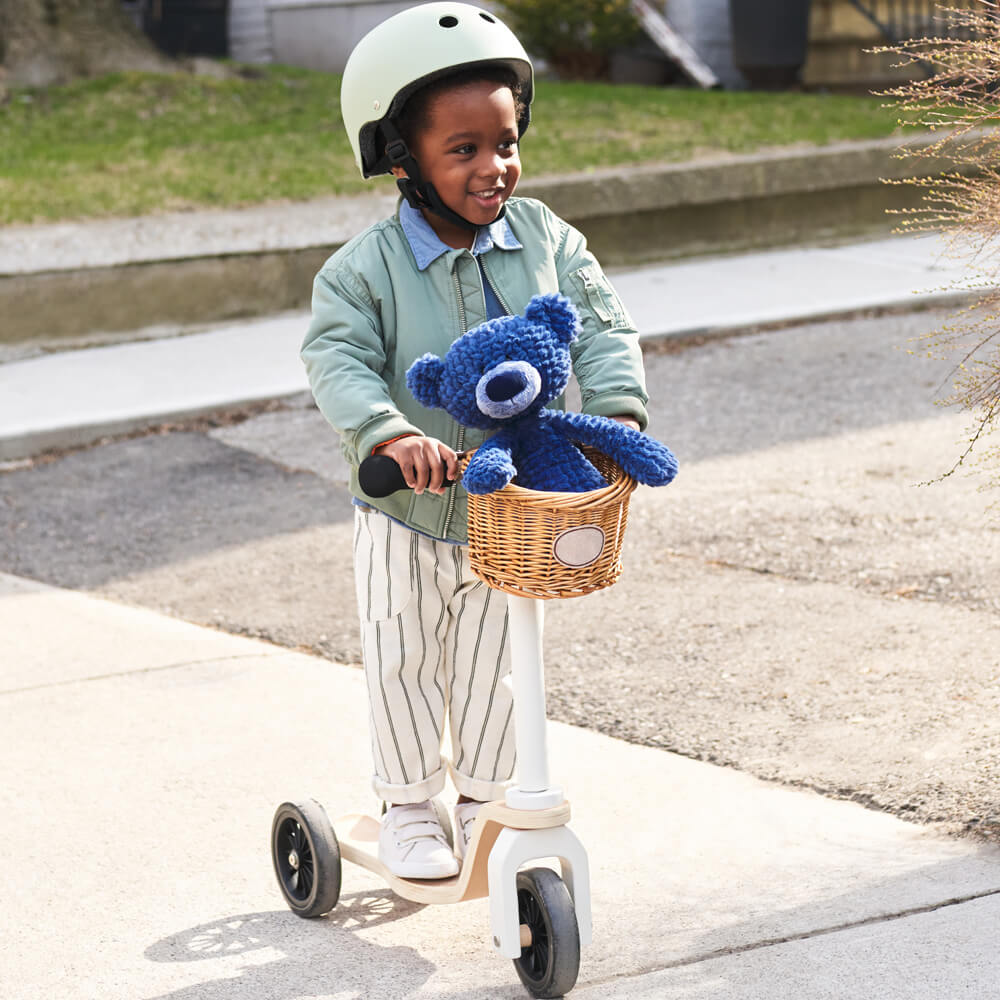 Child riding a scooter with a basket containing a blue toy, wearing a helmet.