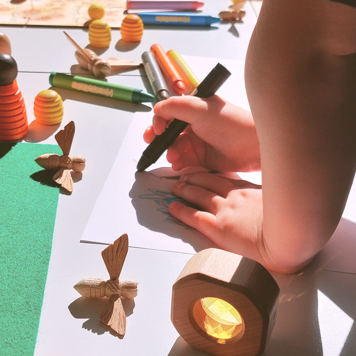Child's hands holding a black crayon with craft materials on a table.