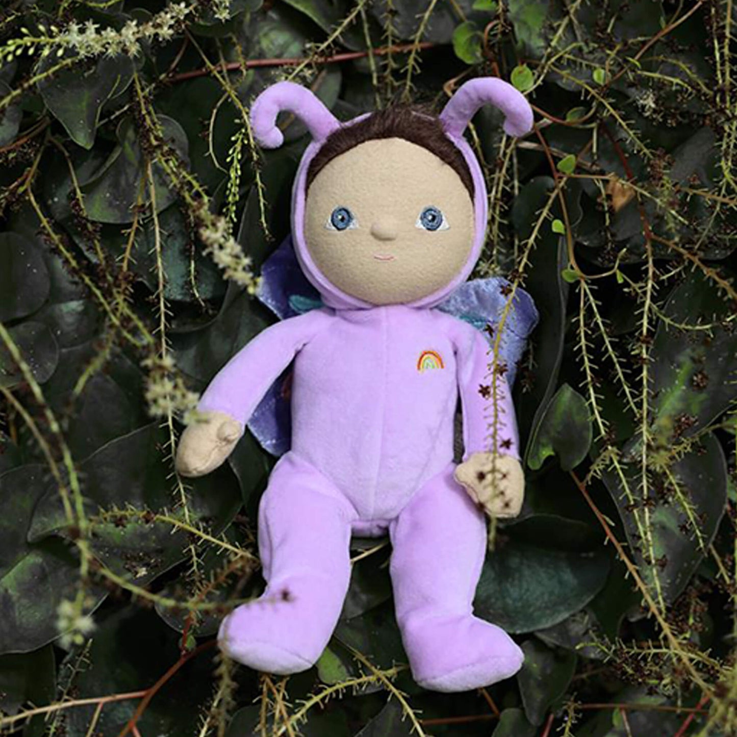 Plush toy dressed in a purple outfit with butterfly wings and antennae, sitting among green leaves.