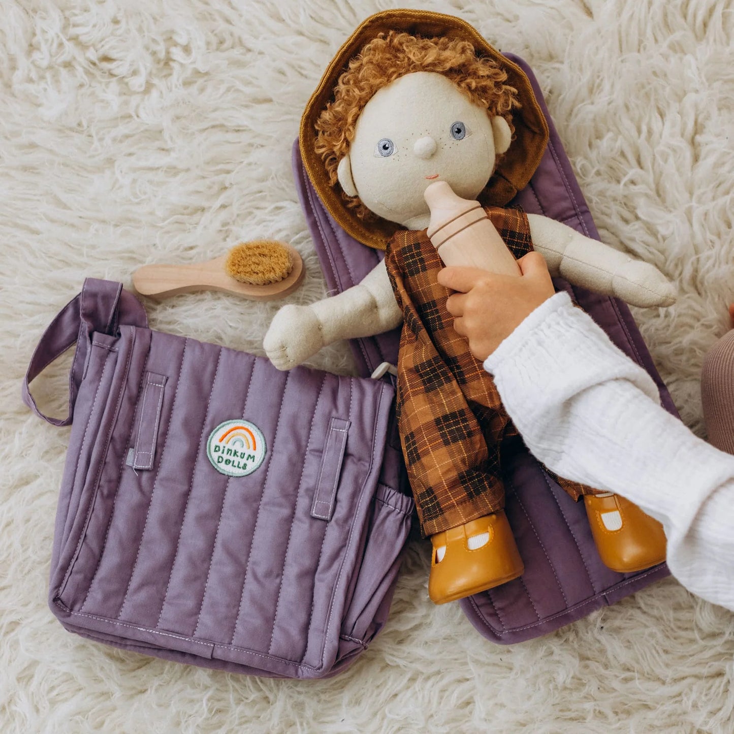Hand holding a doll wrapped in a purple blanket with a brush on a fluffy white surface