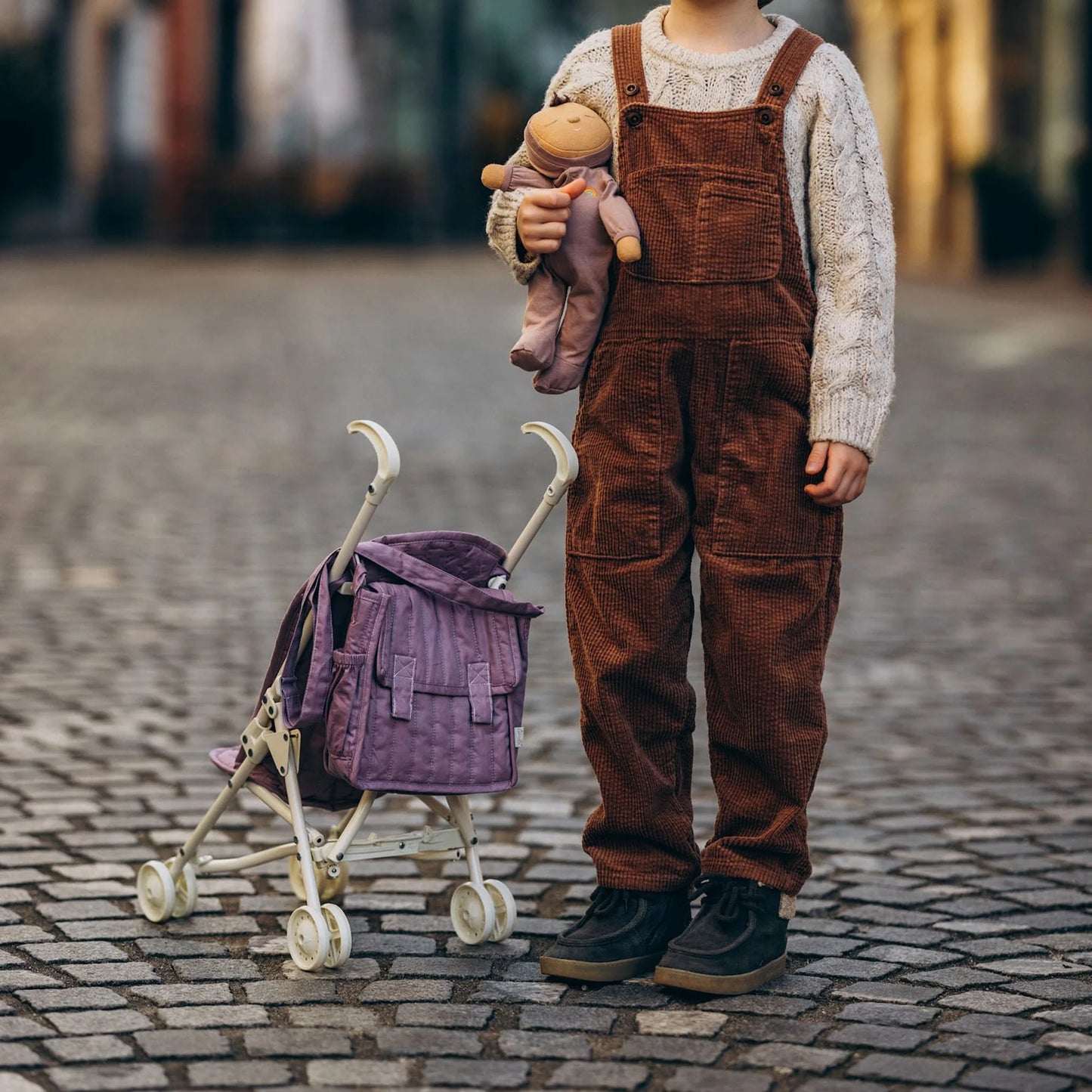 Child in brown overalls holding stuffed animals with a small purple stroller on a cobblestone street.