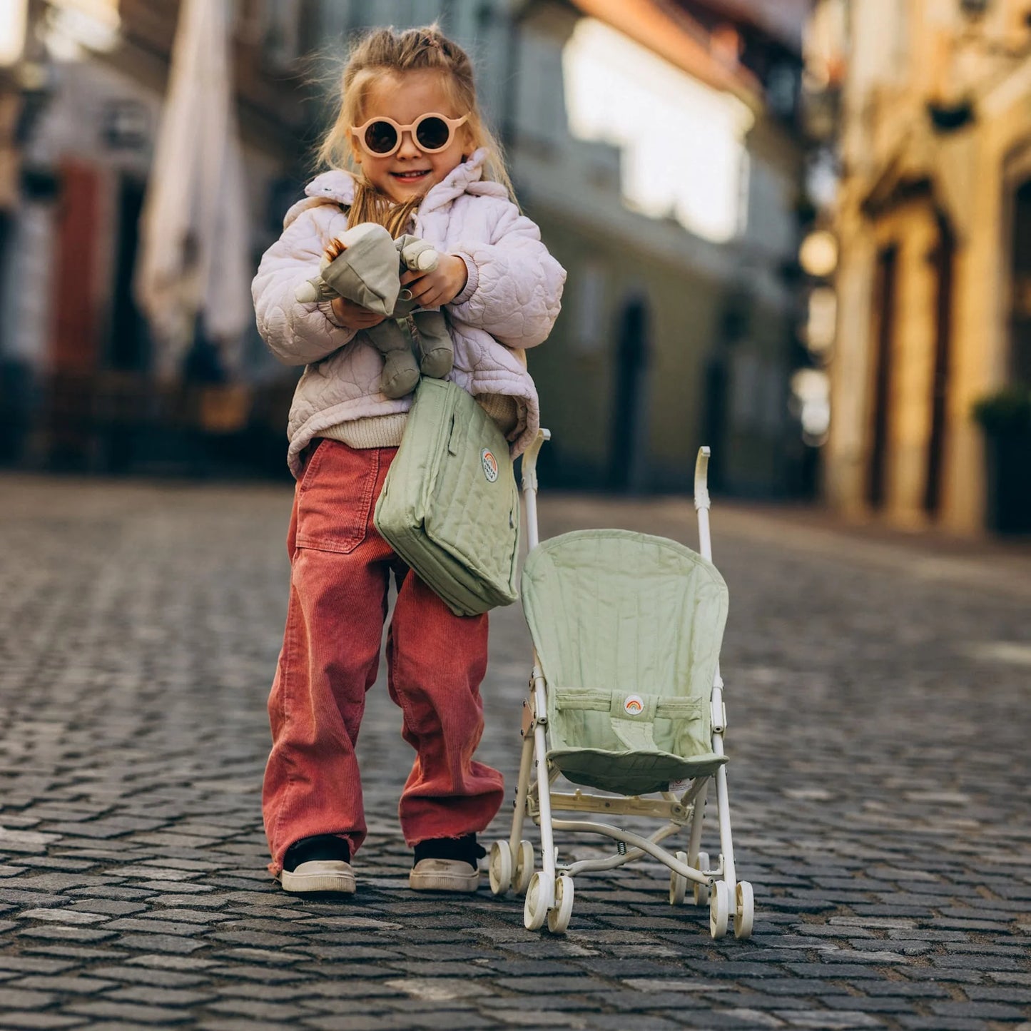 Child holding a green bag and a baby stroller on a cobblestone street.