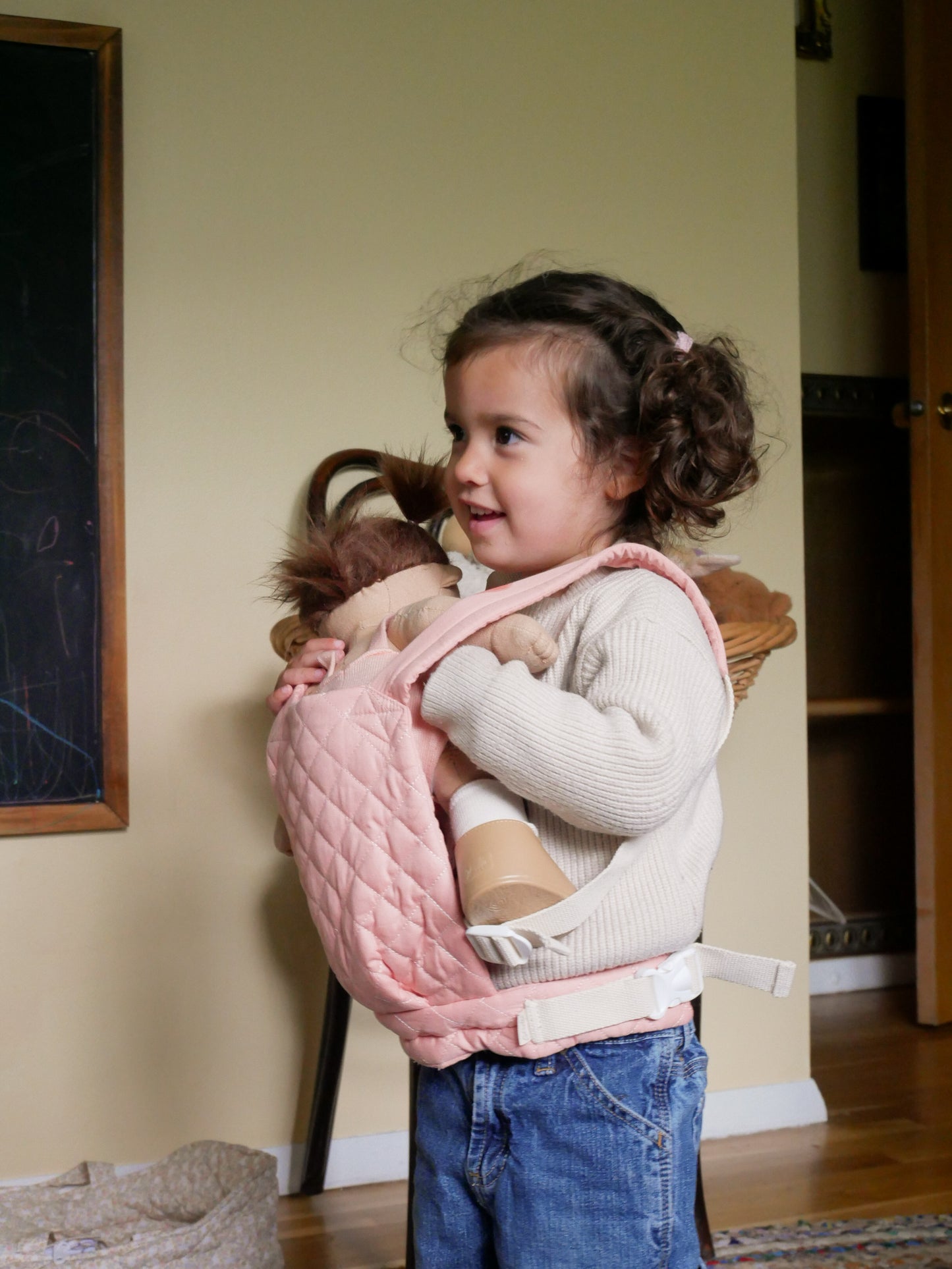 Child holding a doll in a pink carrier with a neutral background