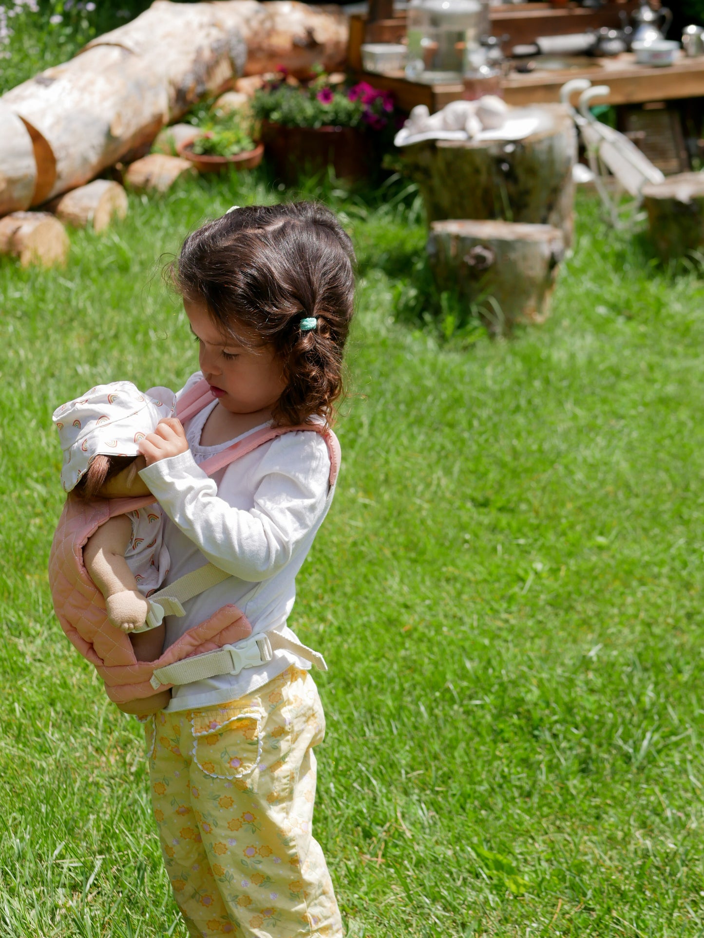 Child holding a doll outdoors on grass with logs and plants in the background