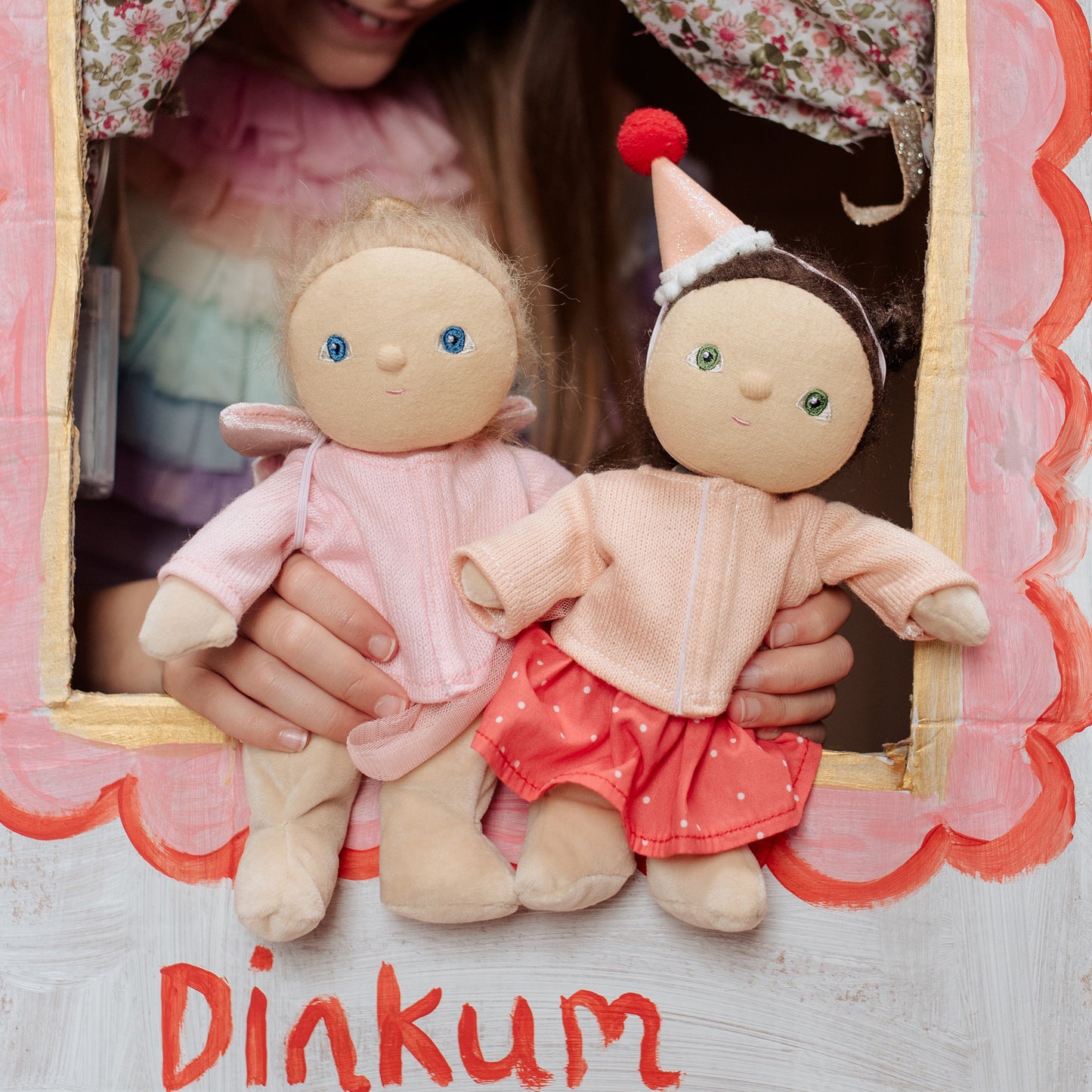 Two dolls held by a person with a 'Dinkum' label, inside a decorative frame.