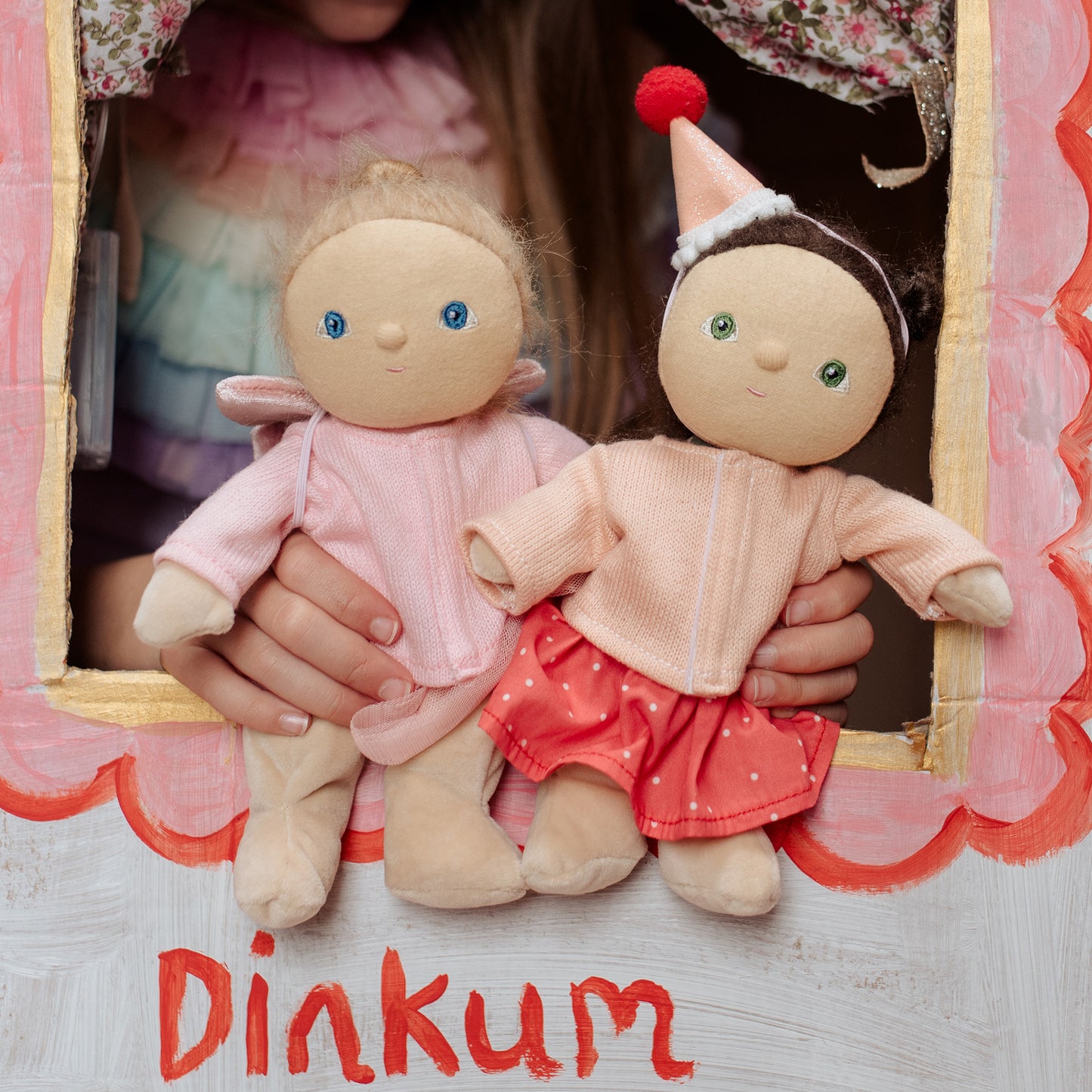 Two dolls held by a hand with a decorative background, featuring the brand 'Dinkum'.