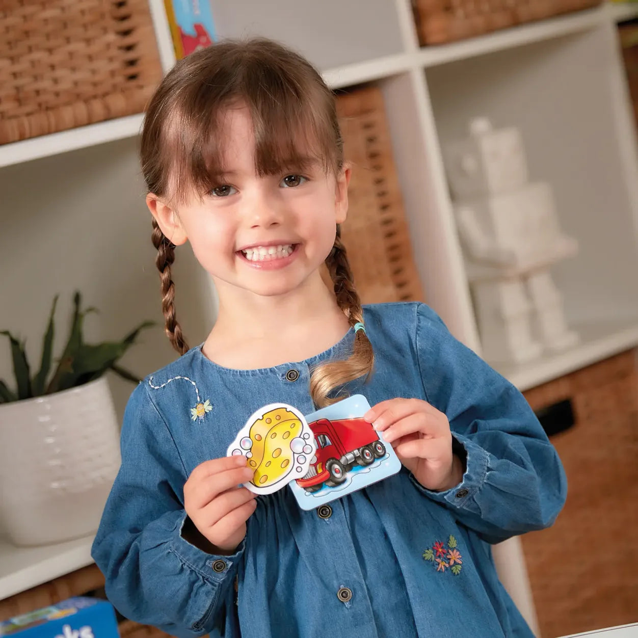 Young girl holding two colorful cards with a red car and yellow sponge design, smiling in a home setting.