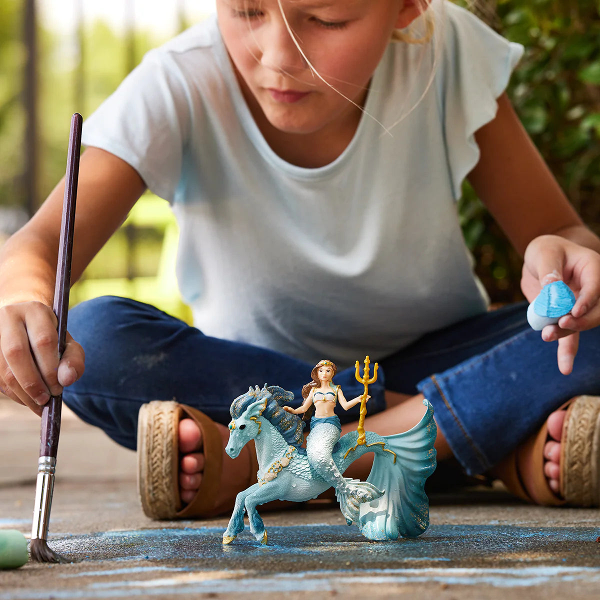 Child painting a figurine of a mermaid and horse on a piece of paper outdoors.