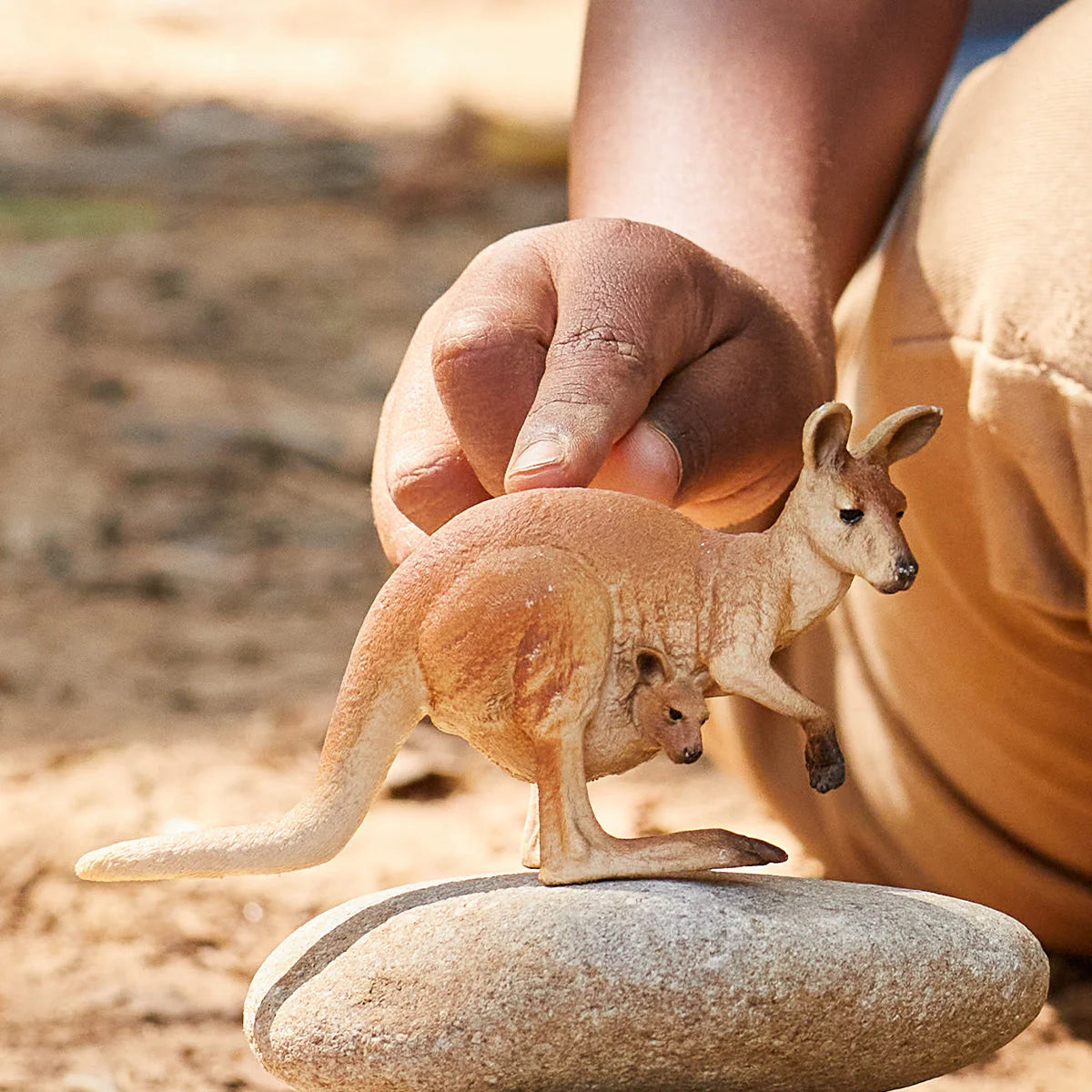 Hand holding a toy kangaroo with a baby on a rock in a natural setting