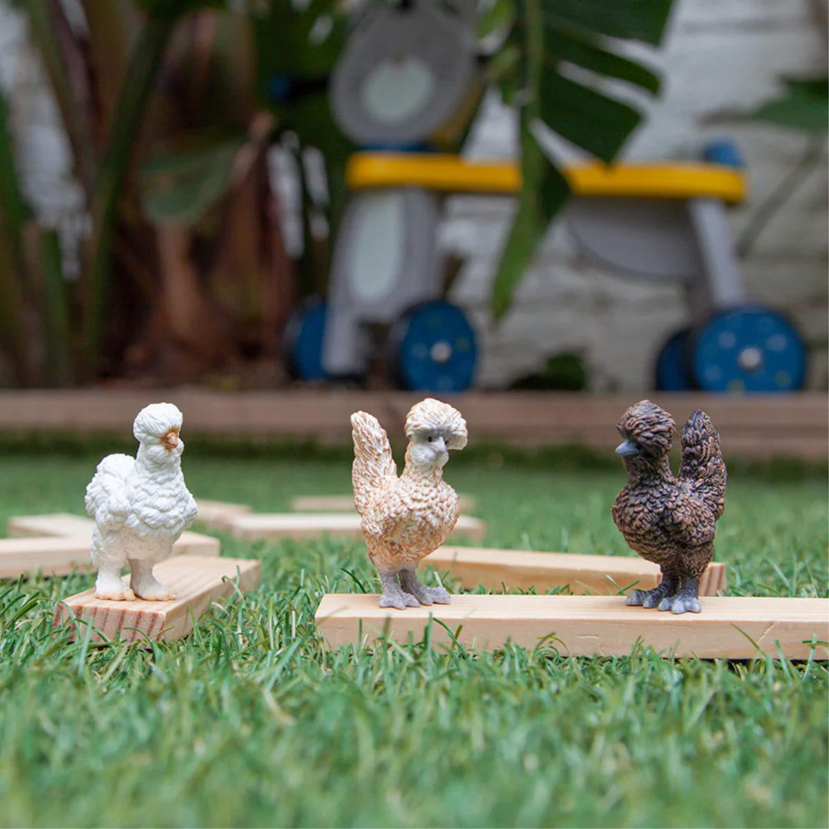 Three toy chickens on wooden stands in a grassy area with a blurred background