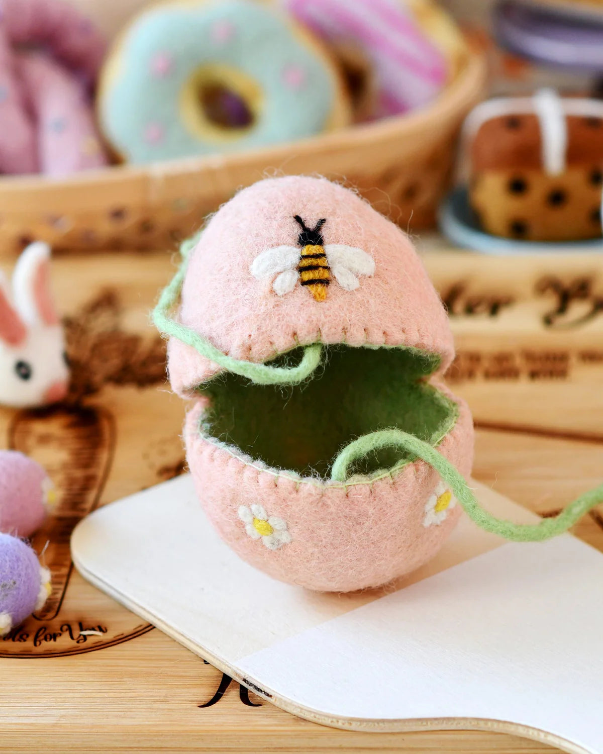 Pink felt egg-shaped container with a bee design on a wooden surface with donuts in the background