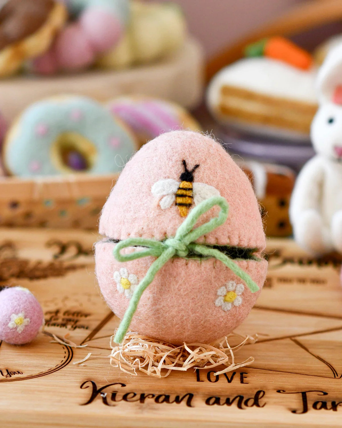 Pink decorative egg with a bee design on a wooden surface with blurred donuts in the background