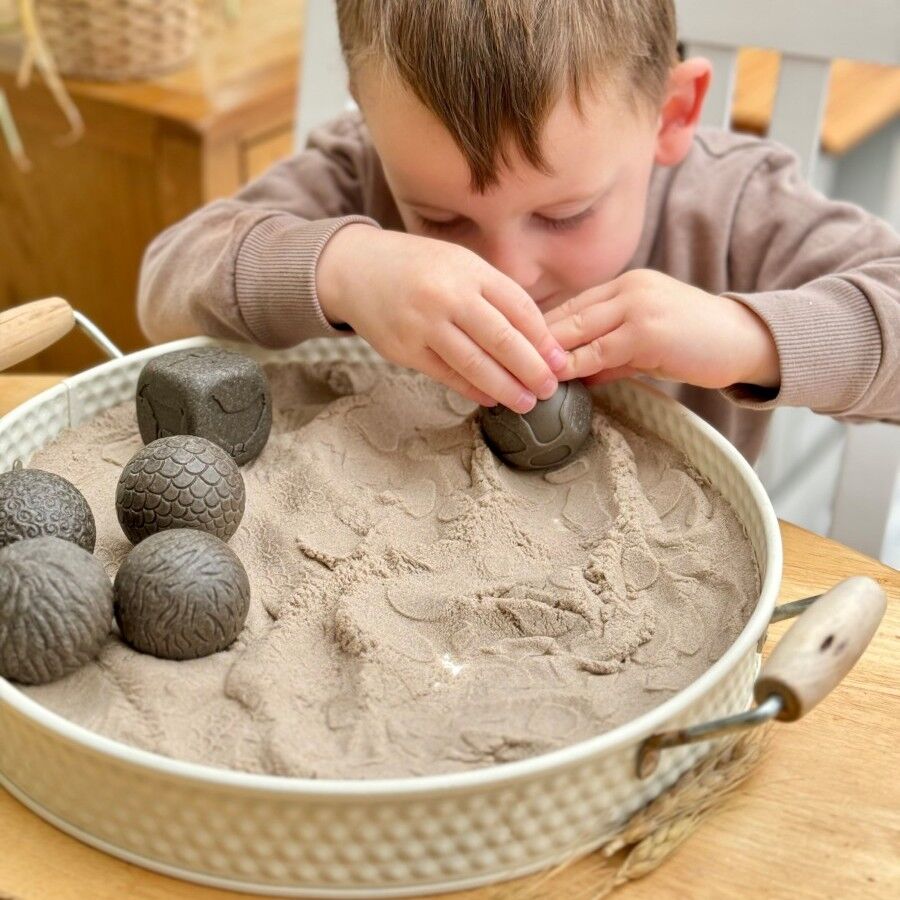 Child playing with sand and rollers in a metal tray on a wooden table.