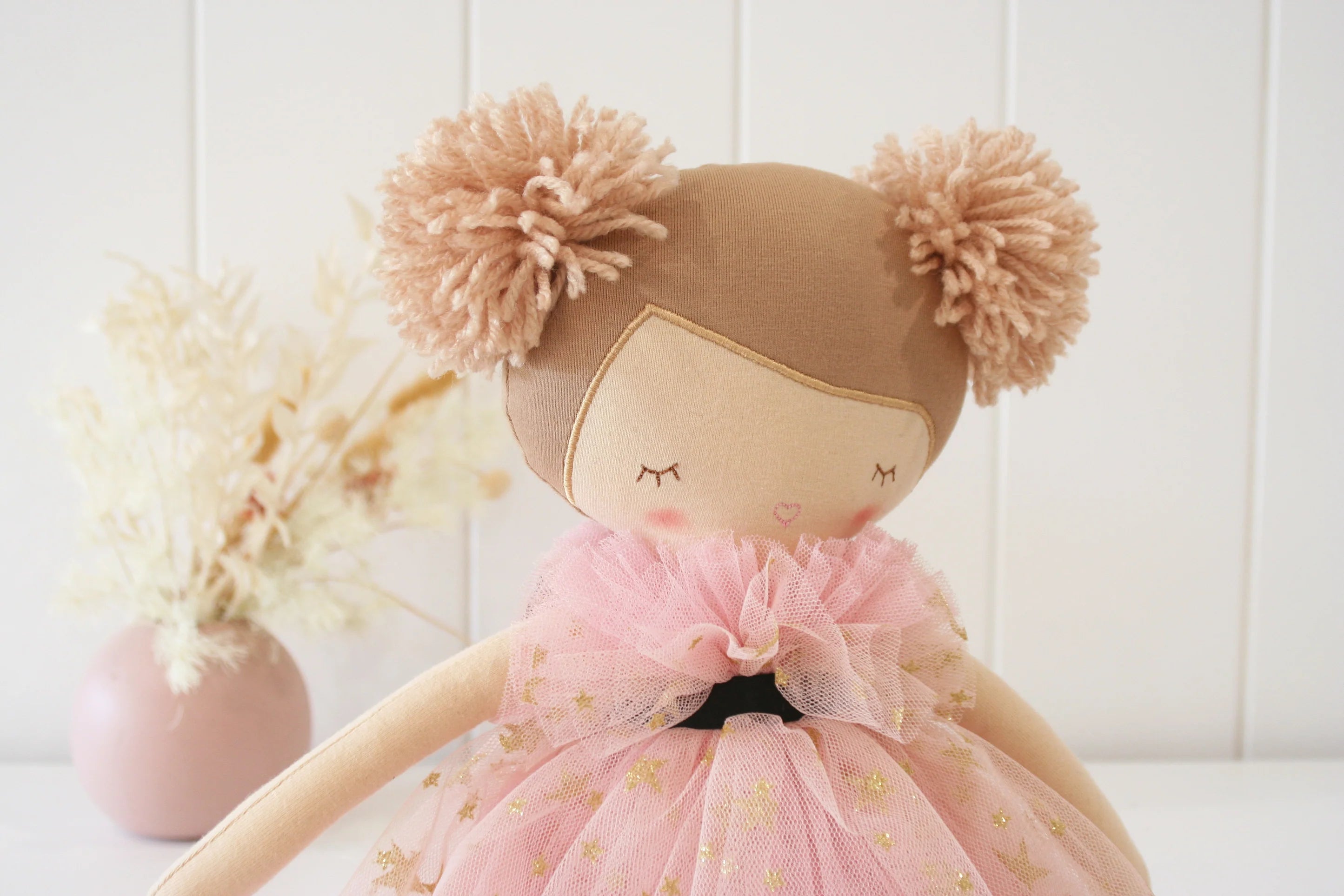 Stuffed doll with brown hair and pink dress in front of a white wall