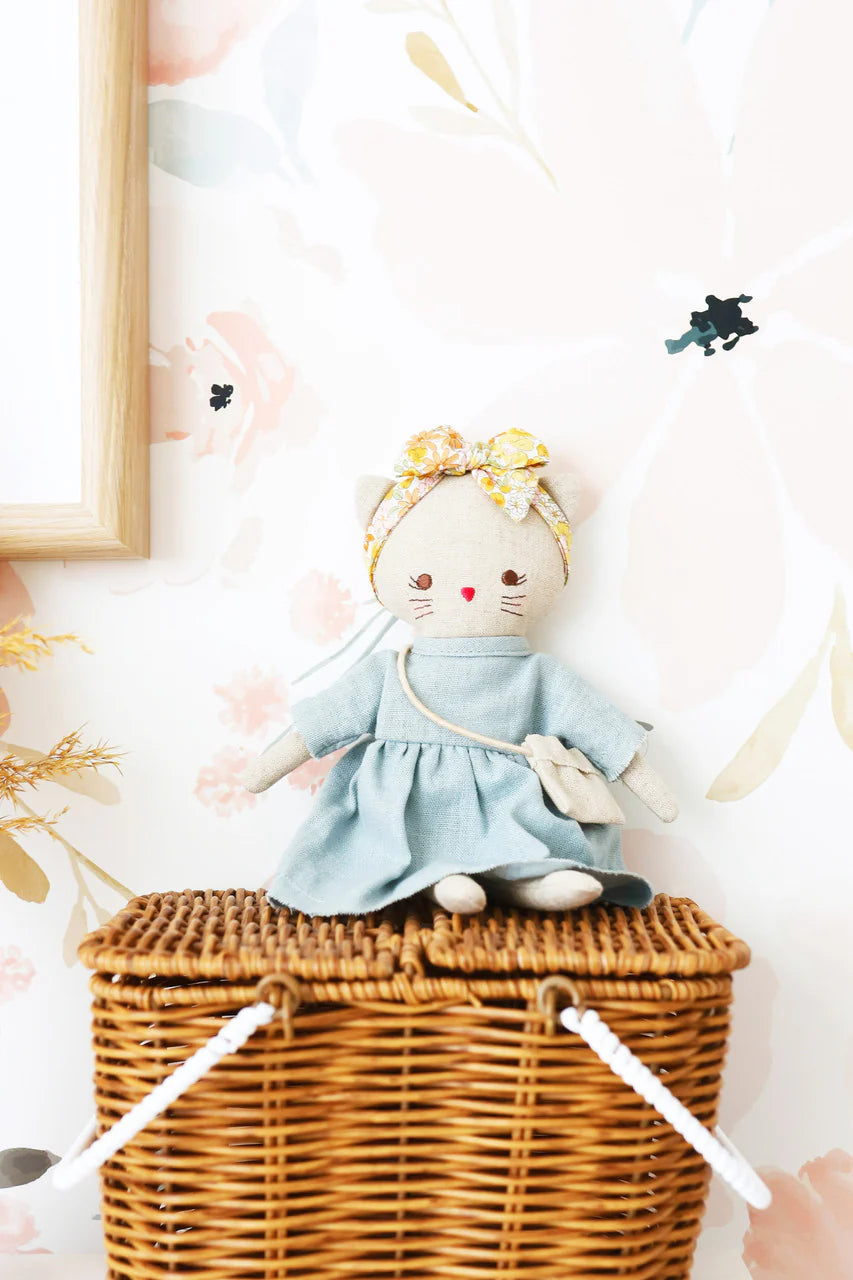 Stuffed toy cat in a blue dress on a wicker basket with a floral wall in the background.