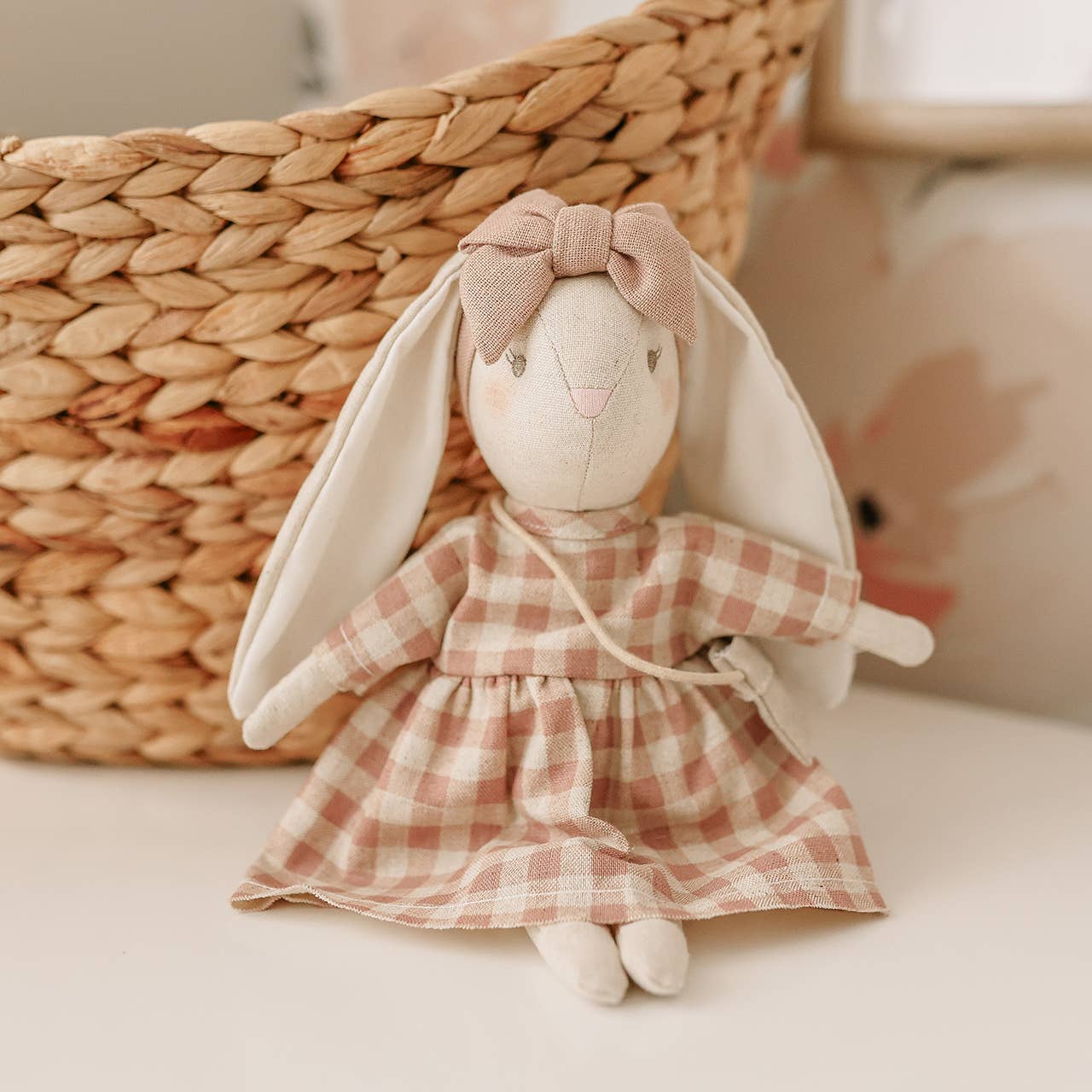 Stuffed rabbit toy with a plaid dress and bow in front of a woven basket.