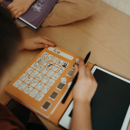 Person using a communication board with symbols and a pen on a wooden table.