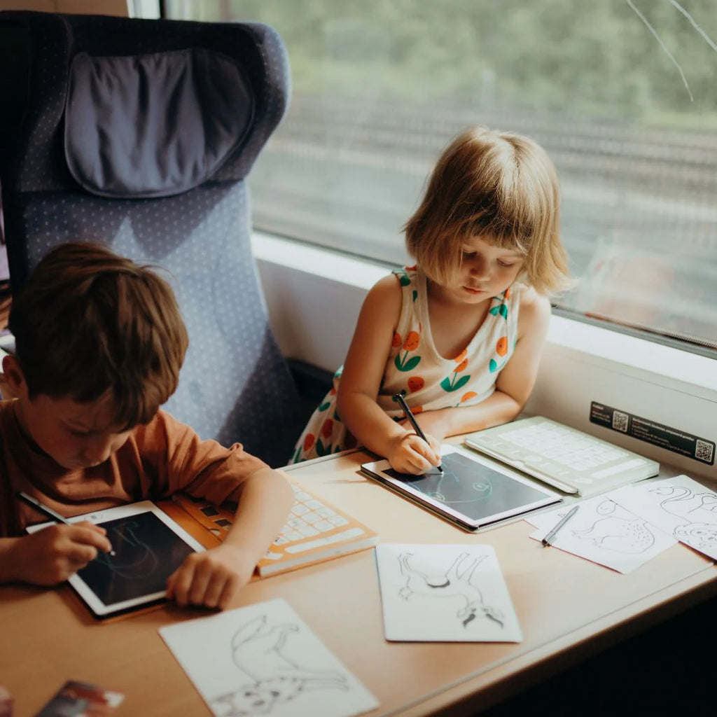 Two children sitting at a table by a window, drawing and writing.