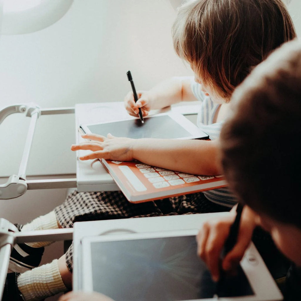 Two children sitting at a desk with one using a tablet and the other drawing on paper.