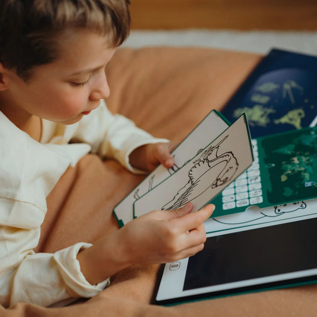 Child holding a coloring book with a tablet open on a bed