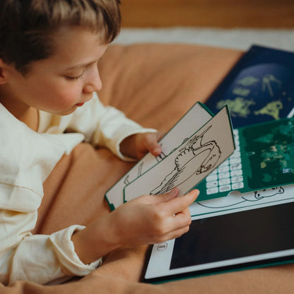 Child holding a coloring book with a tablet open on a bed