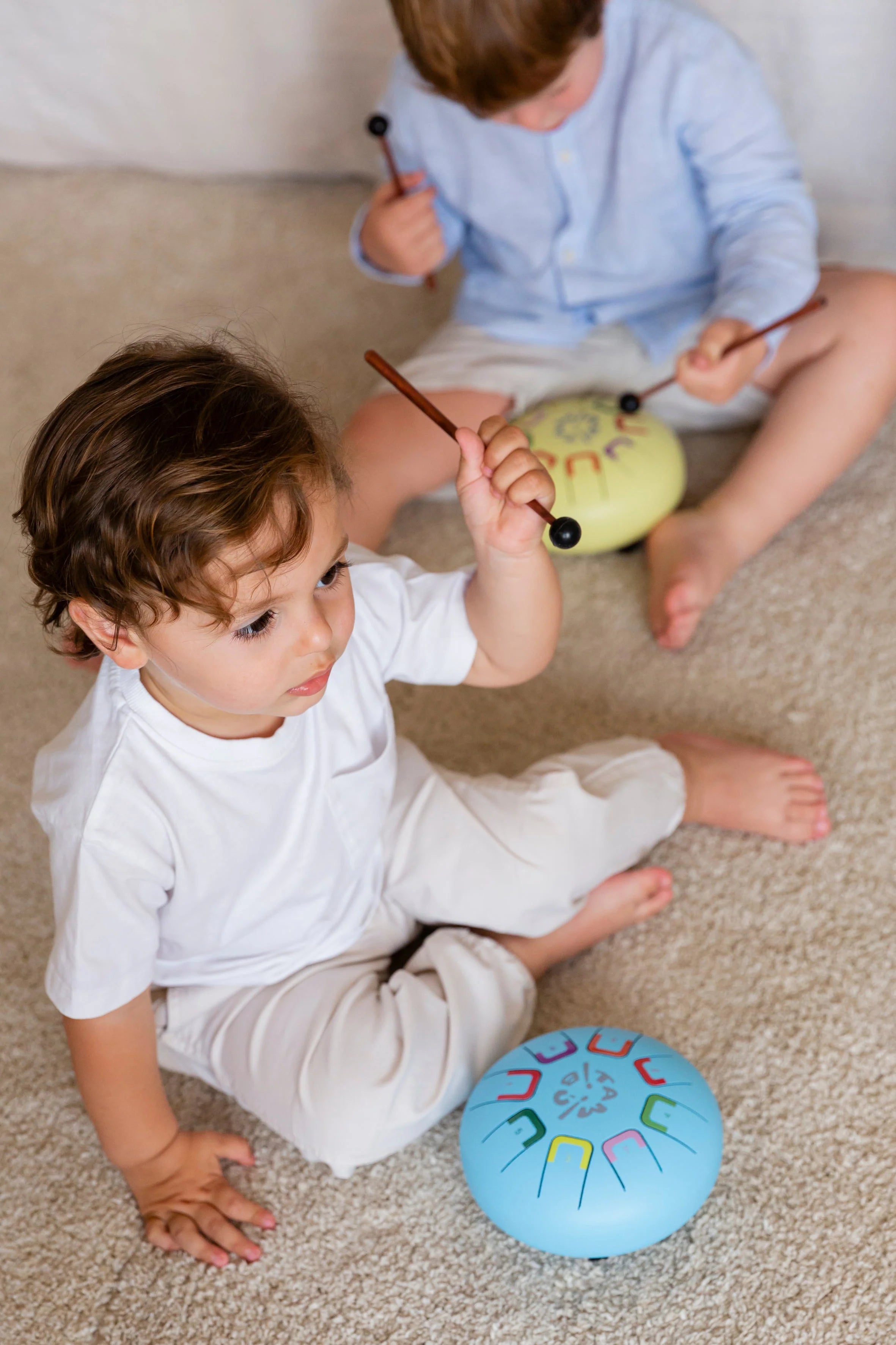 Two children playing with colorful drums on a carpeted floor.