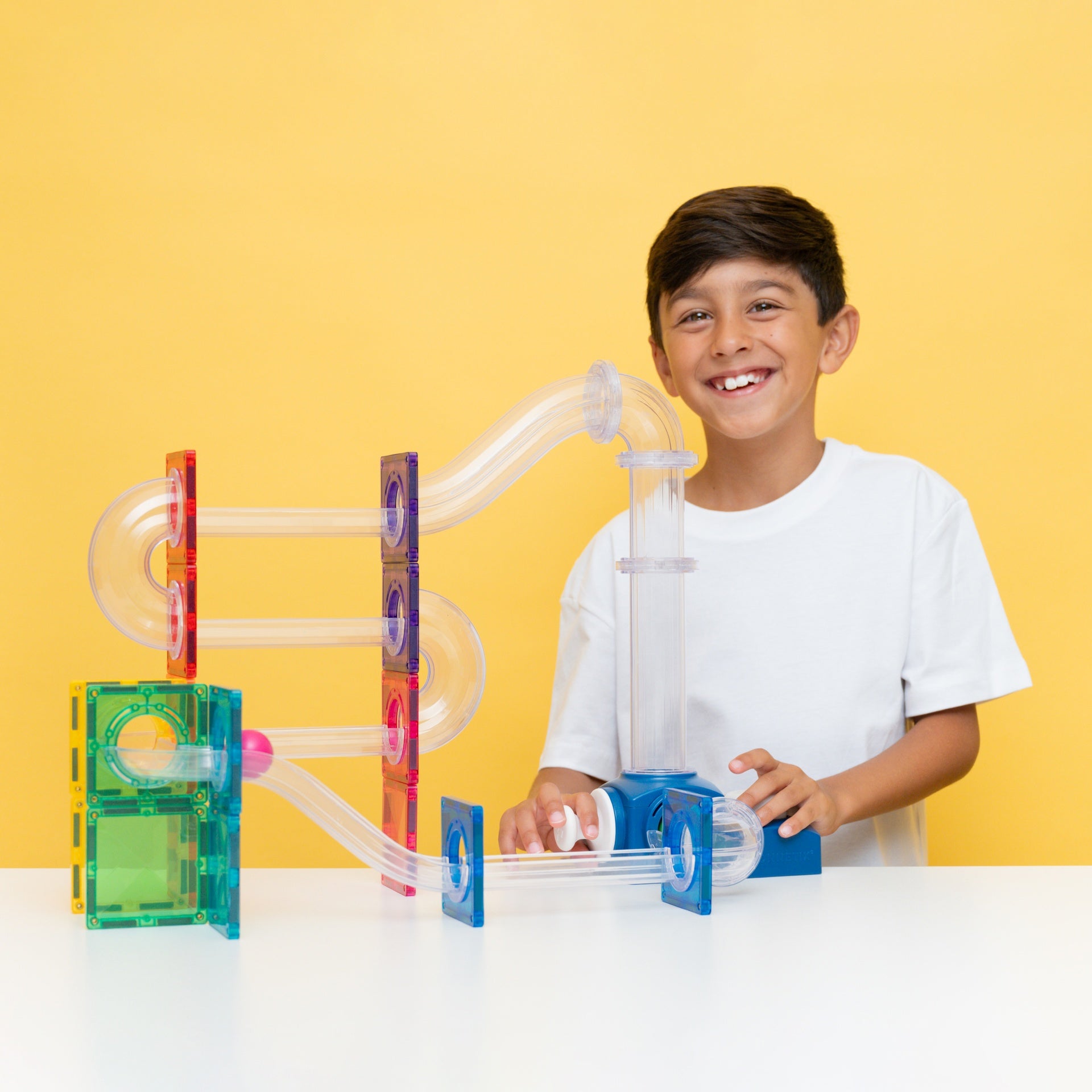 A smiling young boy in a white t-shirt stands behind a colorful magnetic marble run toy set on a white table against a solid yellow background.