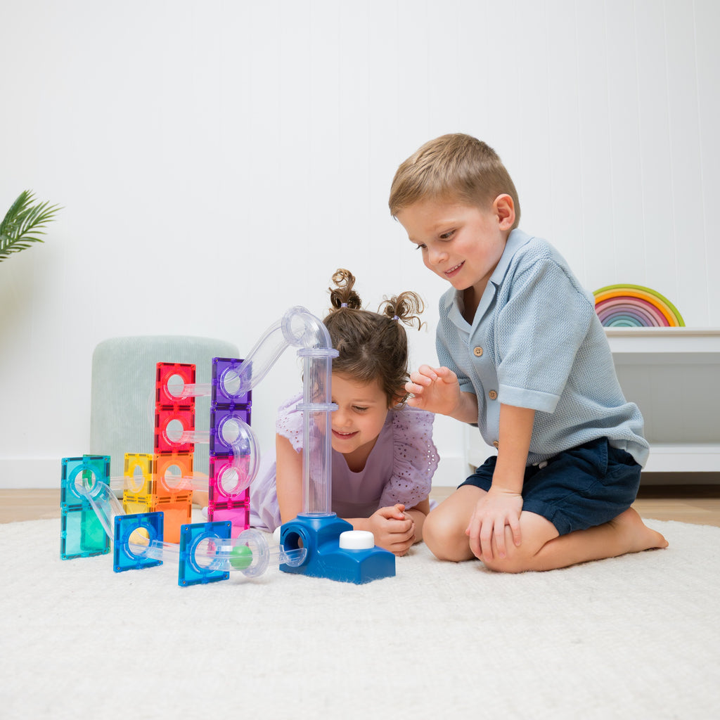 Two children playing with colorful translucent marble-run style blocks and tubes on a white rug in a bright indoor room.