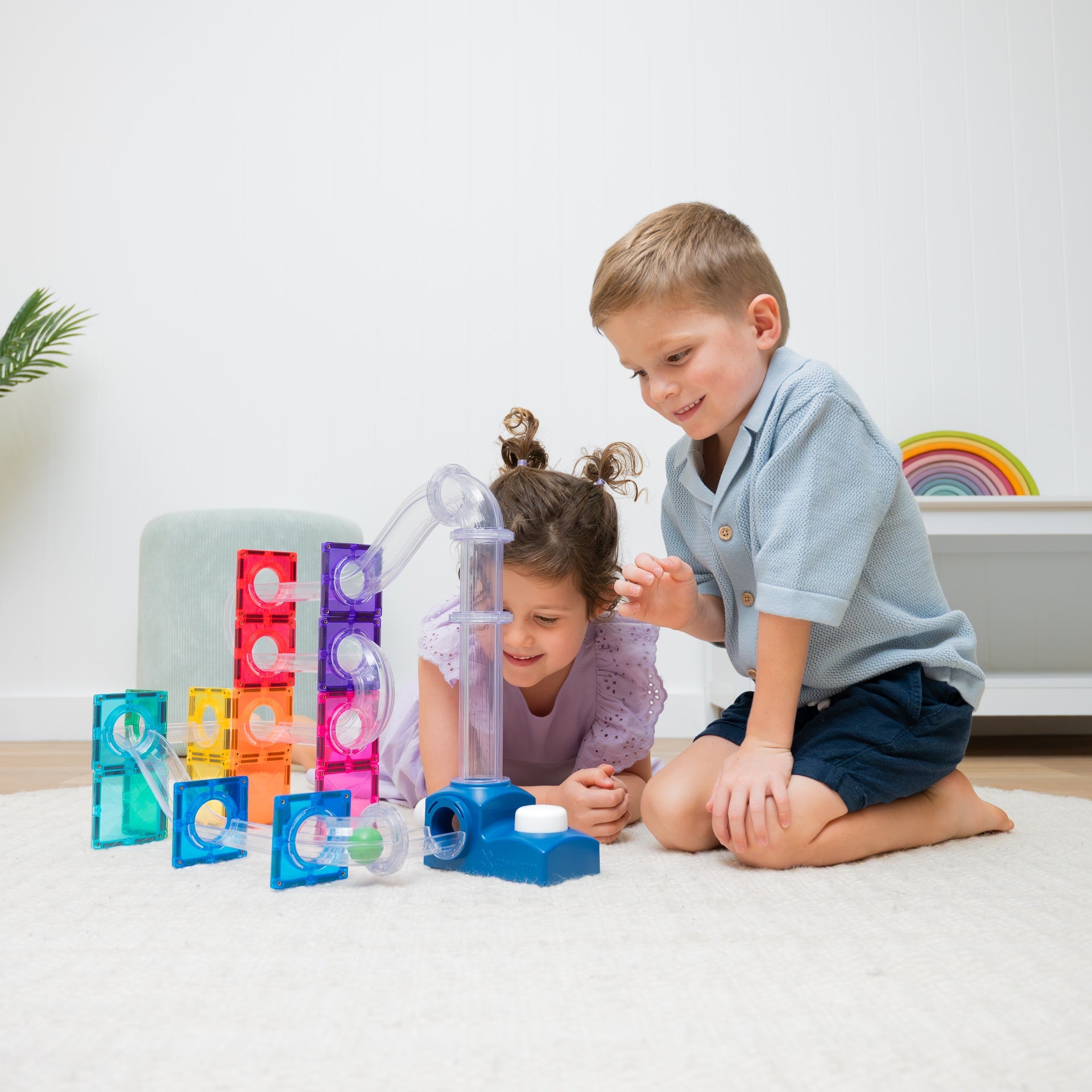 Two children playing with colorful translucent marble-run style blocks and tubes on a white rug in a bright indoor room.