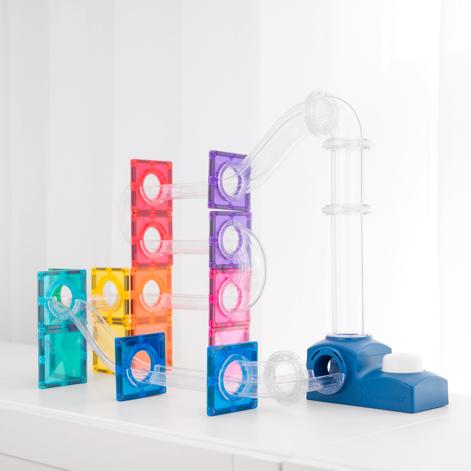 A colorful transparent plastic marble run toy set on a white surface against a white background. The set includes translucent square blocks in teal, yellow, red, orange, purple, and blue, connected by clear curved tubes to a blue base with a clear vertical tube.