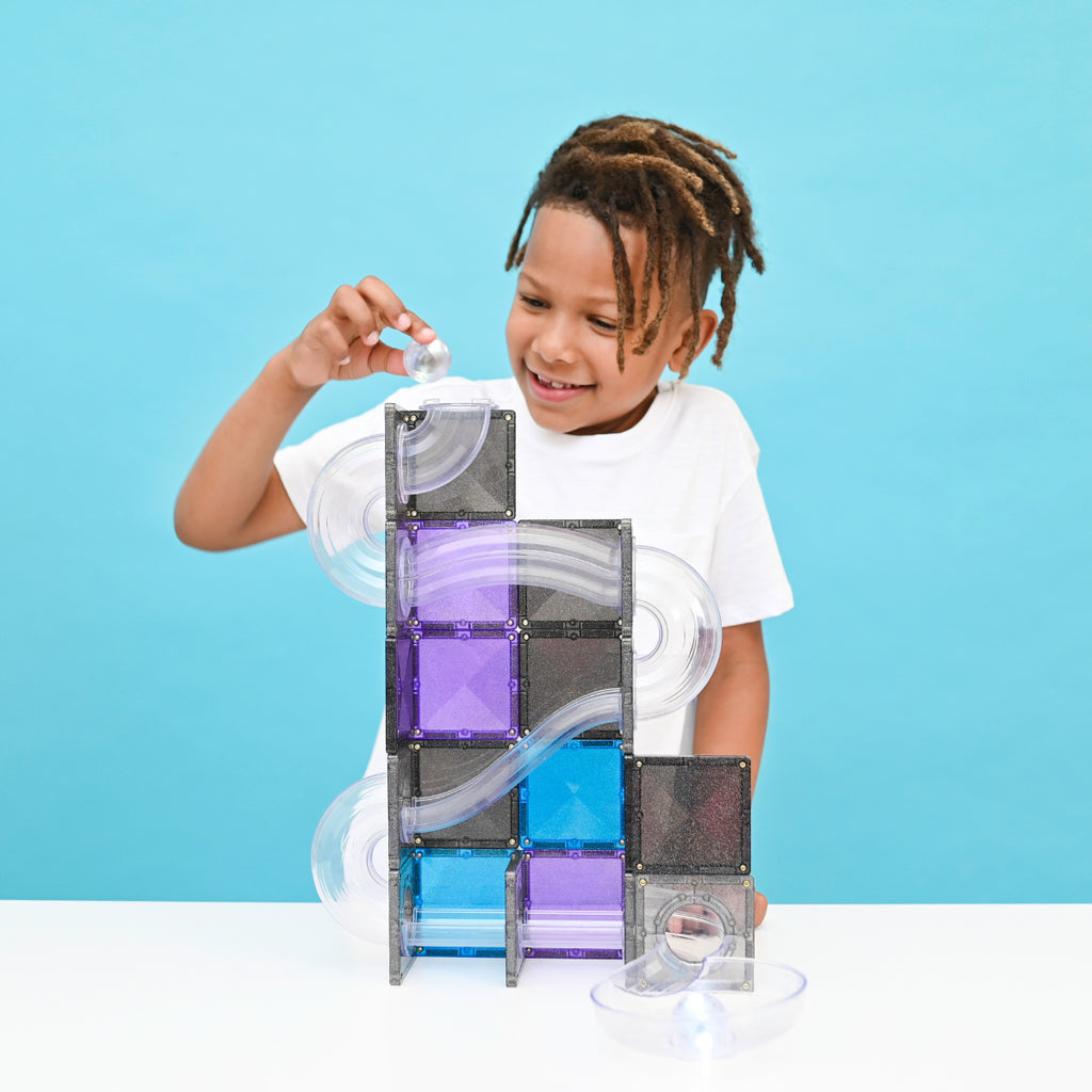 A young boy with dreadlocks smiling and building a clear magnetic marble run tower with translucent blocks and purple, blue, and gray panels on a white table against a bright blue background.