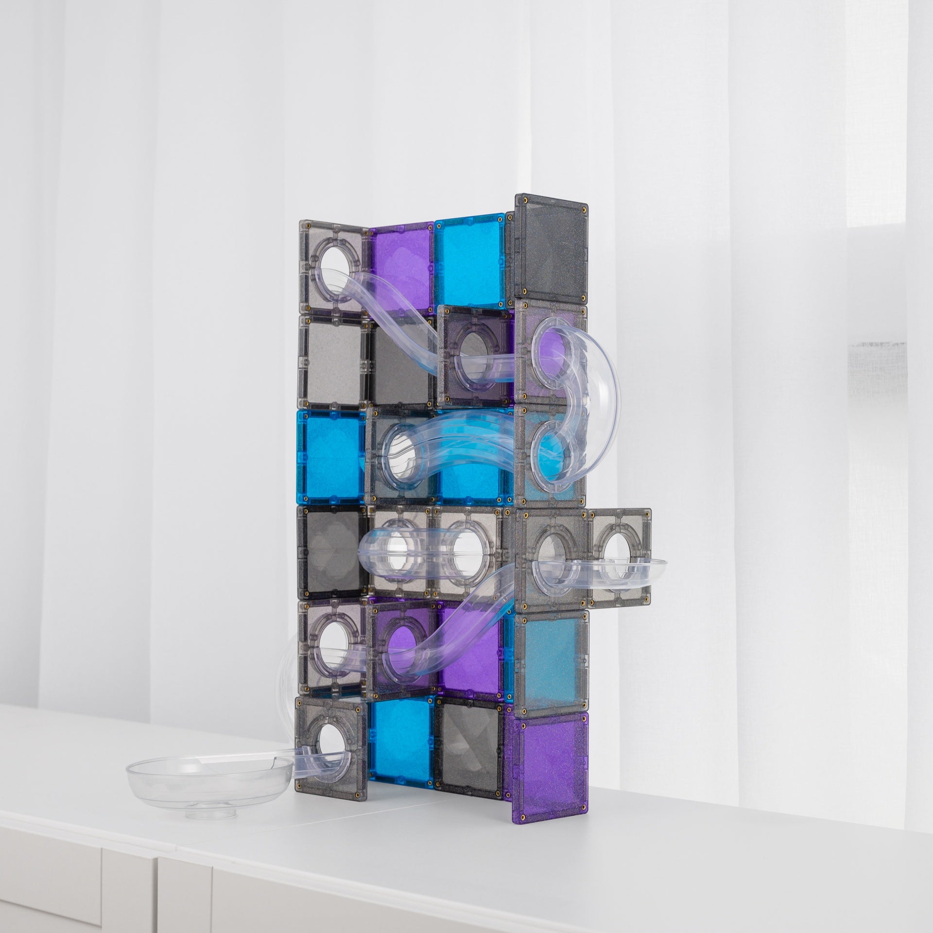 A modular magnetic tile play structure made of translucent gray, purple, and blue square tiles with clear curved tubes, standing on a white surface against a white curtain background.