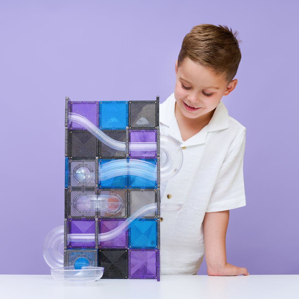 A young boy in a white polo shirt smiles while looking at a magnetic marble run toy built from purple, blue, and black translucent tiles on a white table against a solid lavender background.