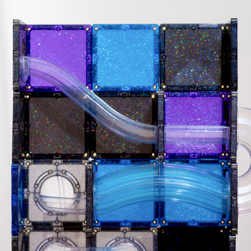A close-up of a transparent block puzzle or magnetic tile assembly with curved clear plastic channels. The tiles are in shades of purple, bright blue, dark glittery black, and silver, arranged in a grid with a black frame.