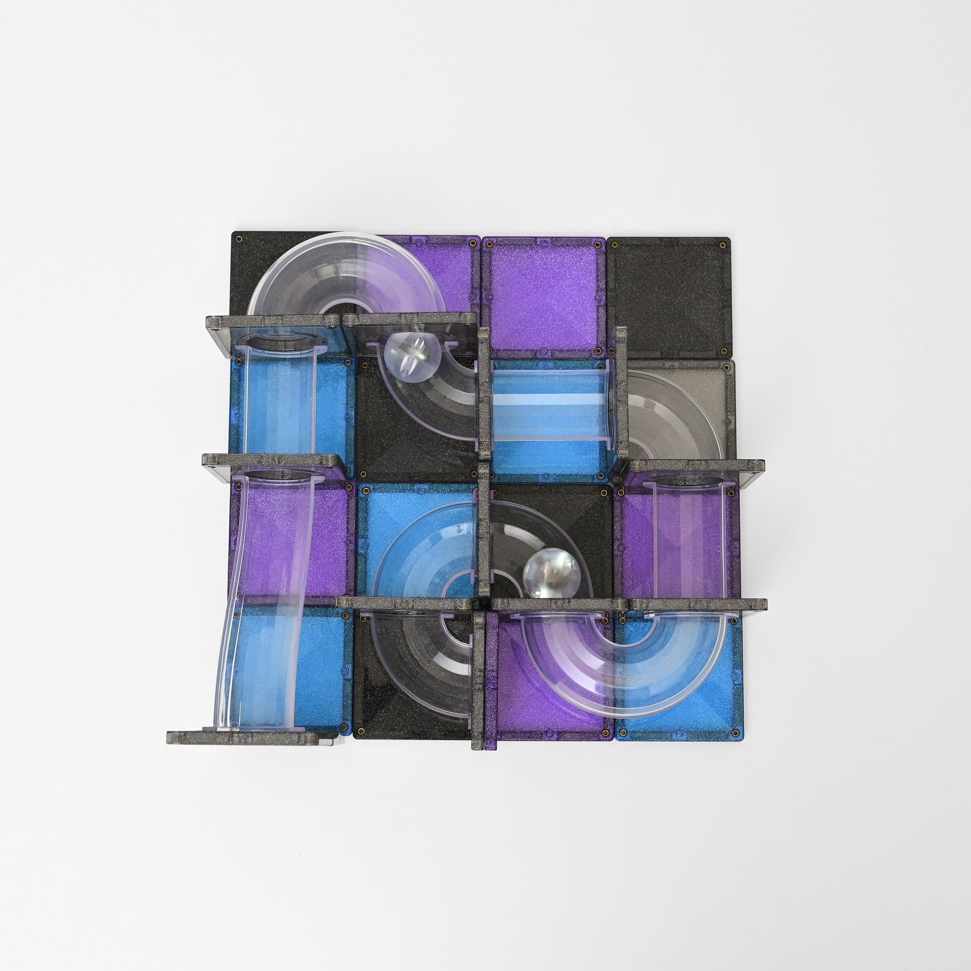 A square magnetic tile toy with a maze-like track, featuring translucent blue and purple tiles, black tiles, clear plastic loops, and a small silver ball on a plain light gray background.