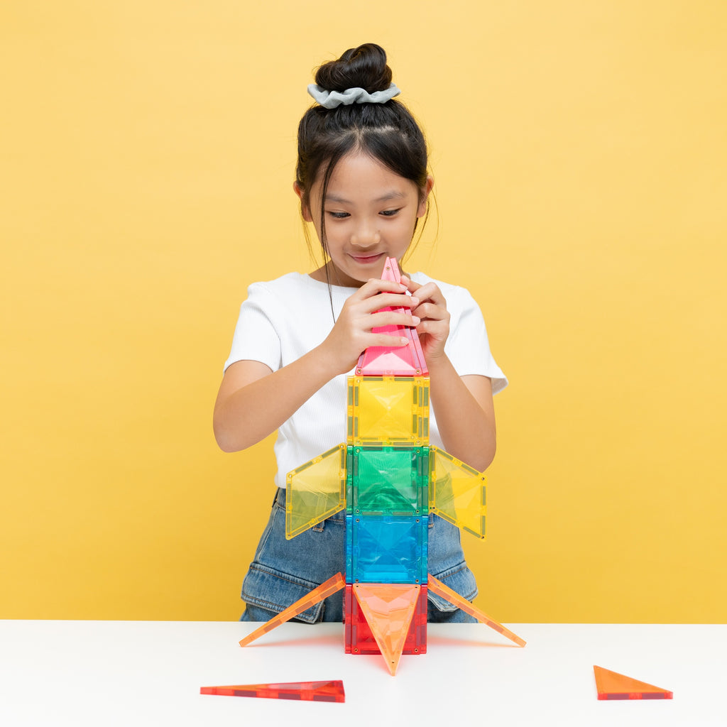 A young girl with her hair in a bun is building a colorful magnetic tile rocket on a white table against a solid yellow background. She is wearing a white t-shirt and denim jeans.