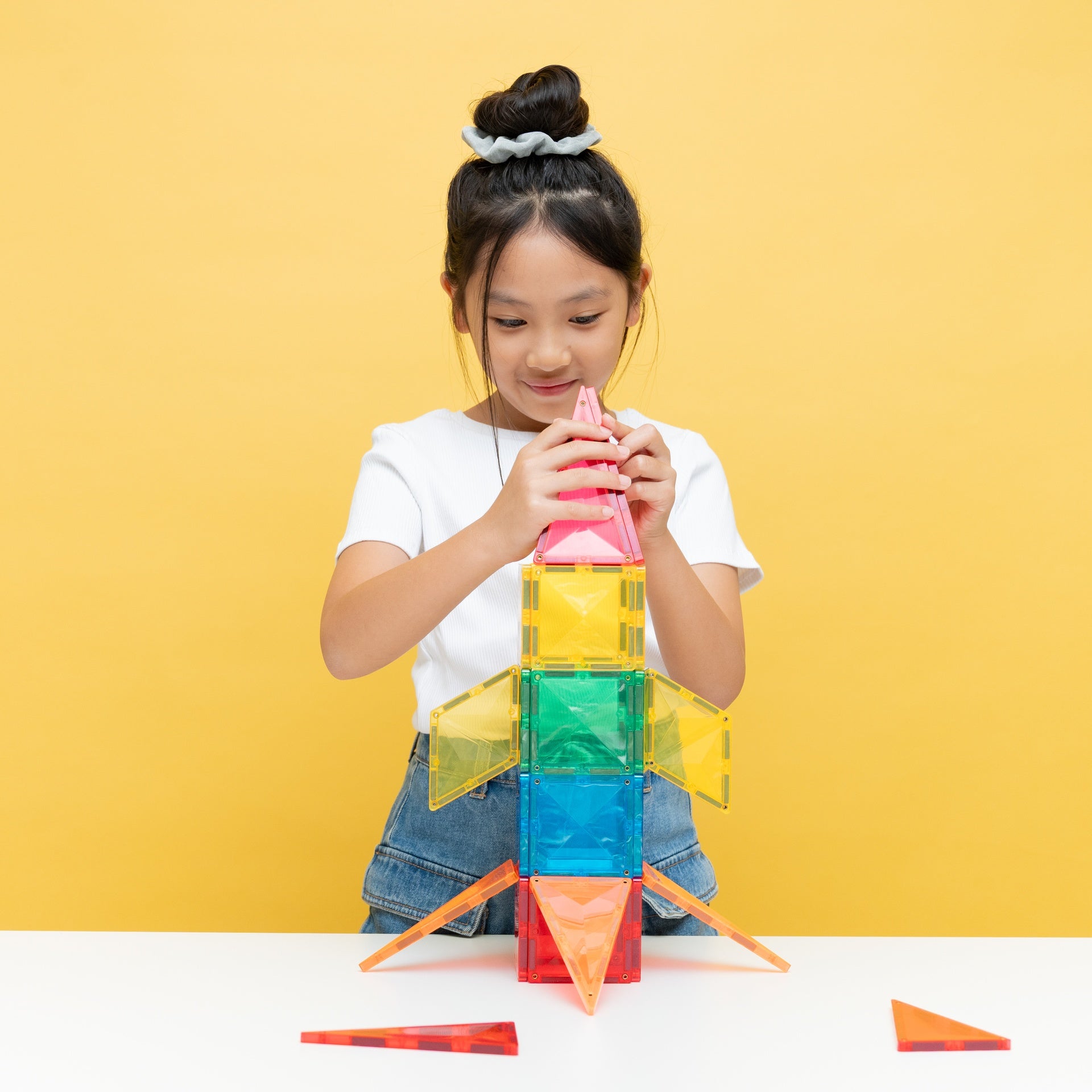 A young girl with her hair in a bun is building a colorful magnetic tile rocket on a white table against a solid yellow background. She is wearing a white t-shirt and denim jeans.
