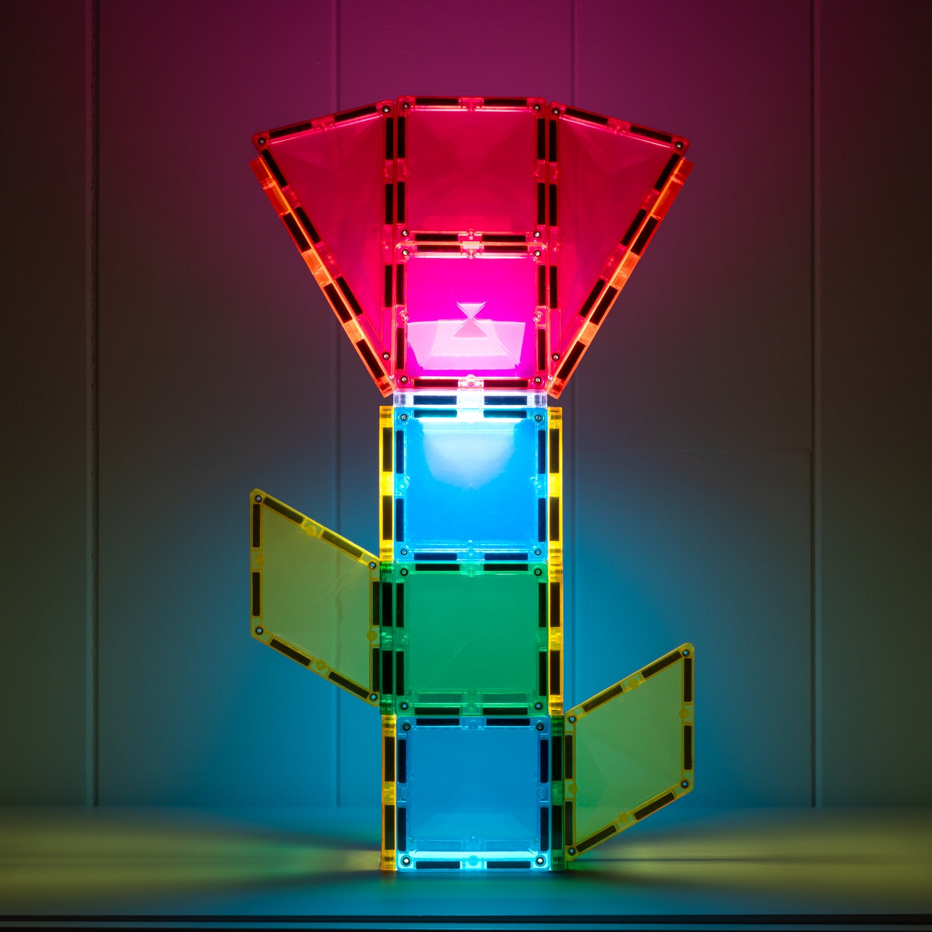 A colorful magnetic tile structure made of translucent acrylic panels with black magnetic connectors, glowing with pink, blue, green, and yellow light against a dark studio background.
