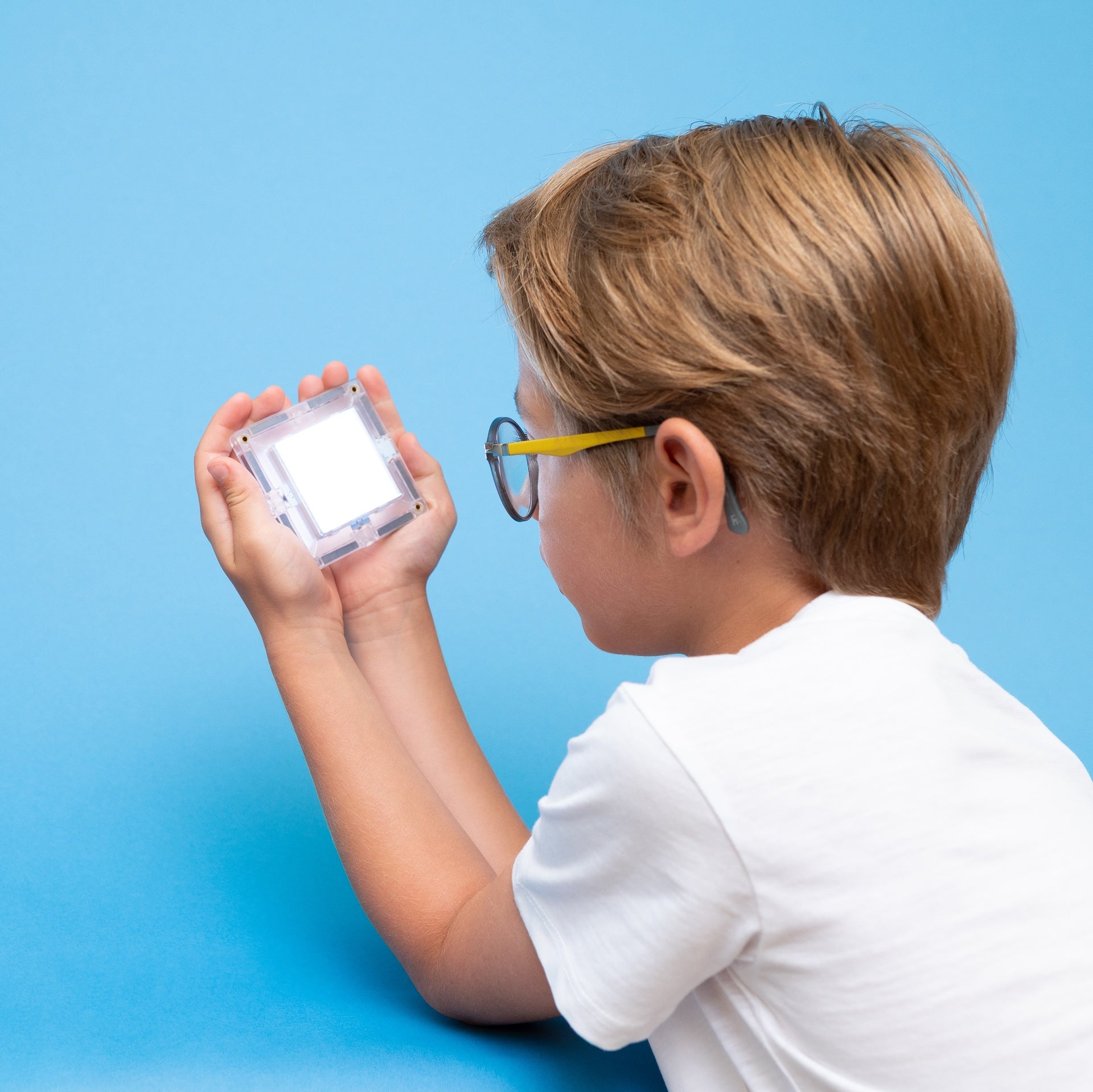 A child wearing a white t-shirt and yellow-framed glasses holds a small clear square object up to their eye against a solid blue background.