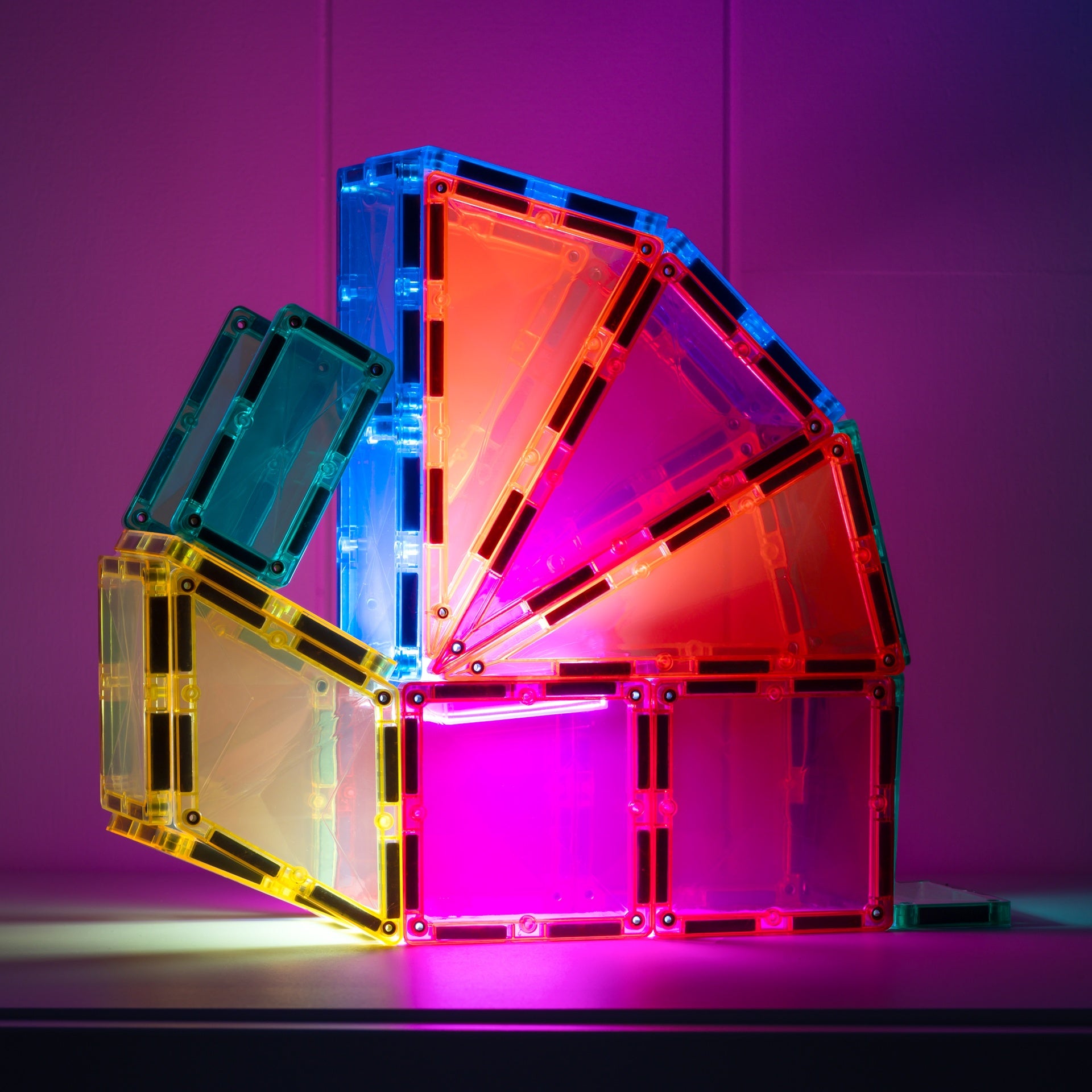 A set of translucent magnetic building tiles arranged in a fan-like structure on a dark surface, glowing with pink, orange, blue, and yellow light against a purple-lit background.