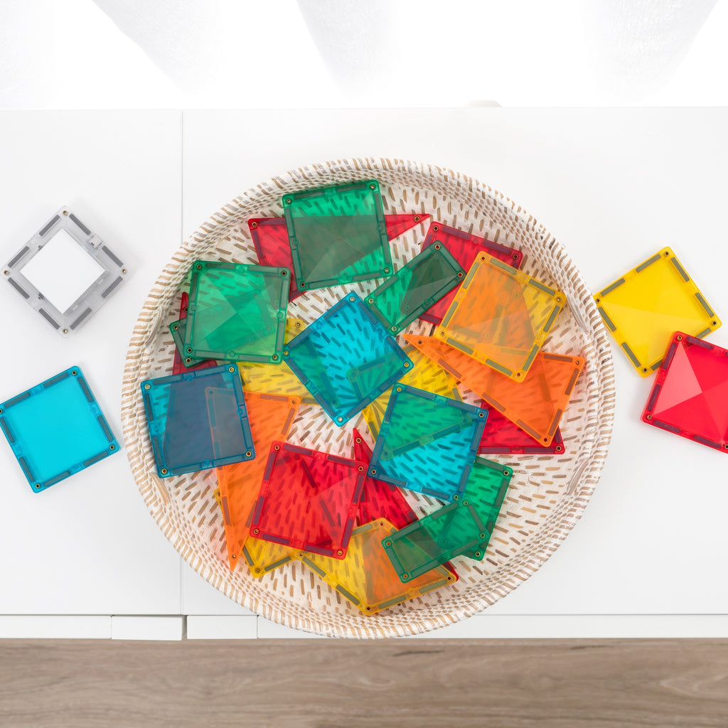 A top-down view of colorful translucent square solar panel tiles arranged in a round woven tray on a white surface, with a few additional tiles placed beside the tray.