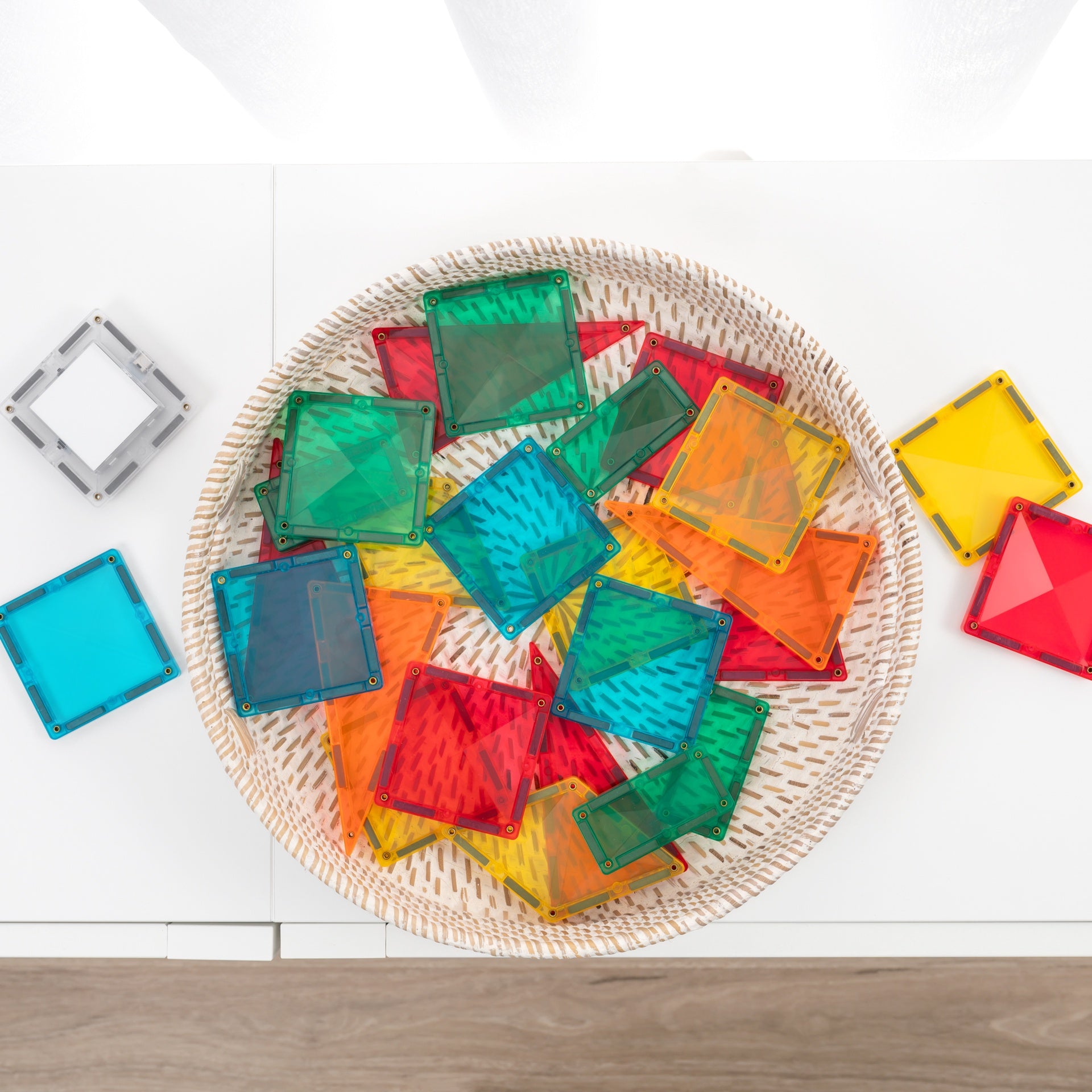 A top-down view of colorful translucent square solar panel tiles arranged in a round woven tray on a white surface, with a few additional tiles placed beside the tray.