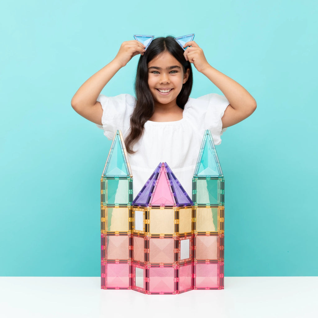 A young girl with long dark hair smiles while adjusting blue cat-ear headphones on her head, standing behind a colorful magnetic building block castle on a white table against a light blue background.