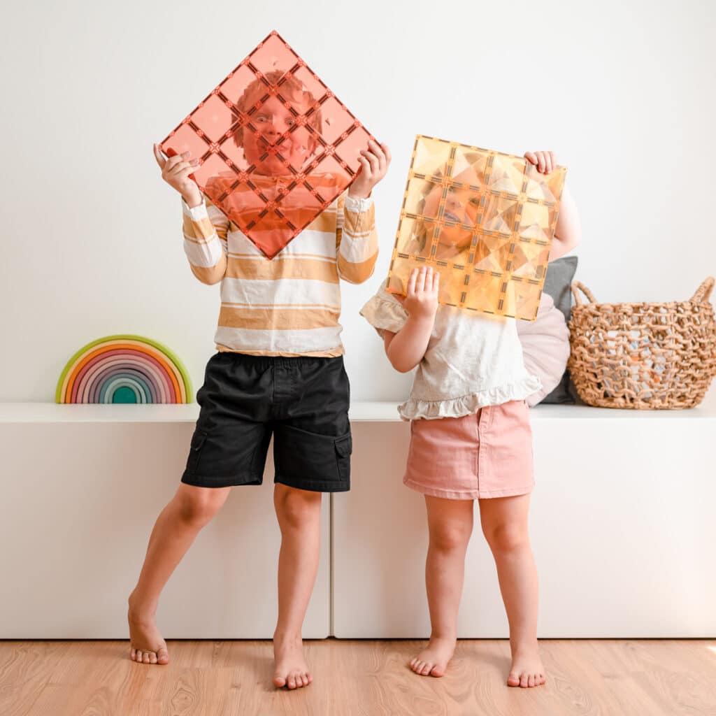 Two children holding colorful geometric shapes against a white wall.